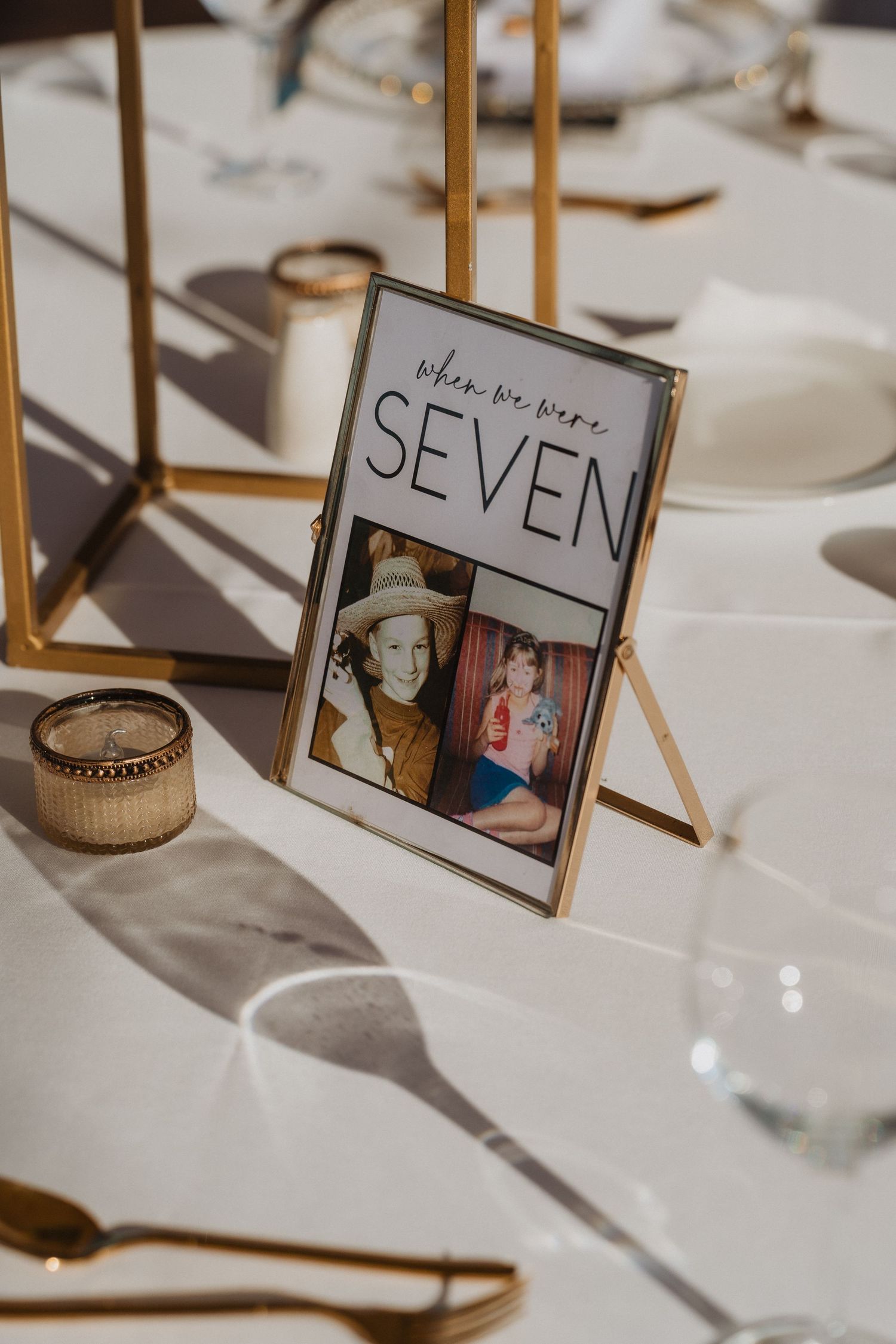 A wedding table number display featuring photos in a gold frame with 'SEVEN' text, set among place settings and decor.