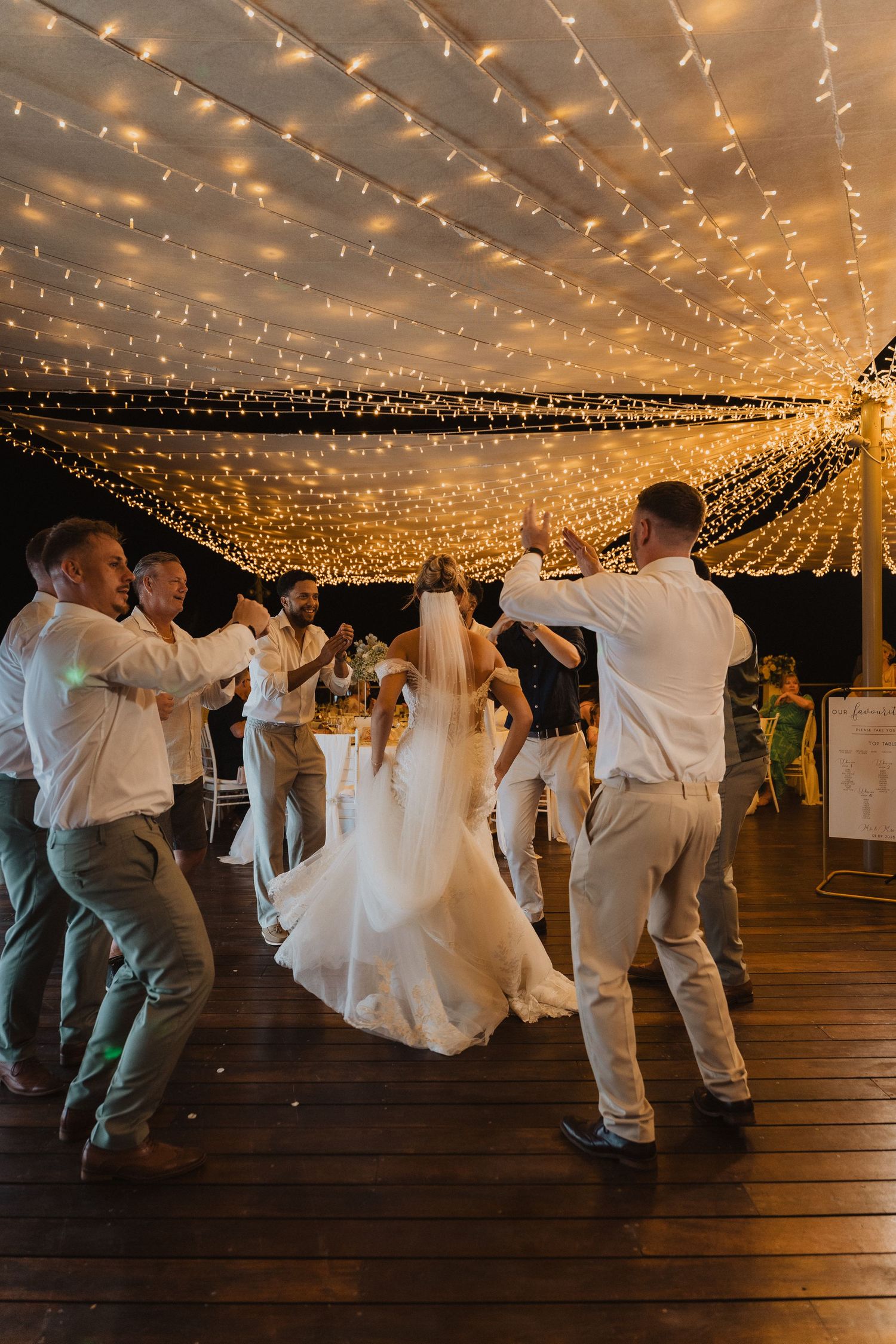 Wedding guests dance on a wooden deck under a magical canopy of twinkling string lights at an outdoor evening reception.