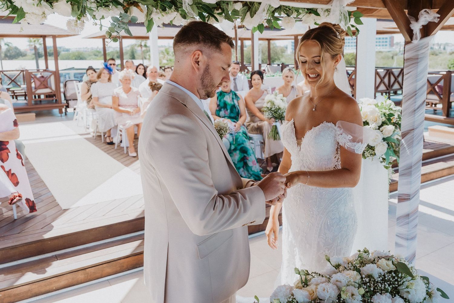 A romantic outdoor wedding ceremony with a couple exchanging rings under a decorated canopy on a sunny day.