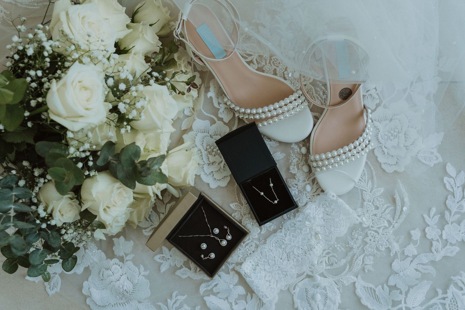 Wedding details with white roses bouquet, elegant bridal shoes, and jewelry boxes on delicate lace fabric.