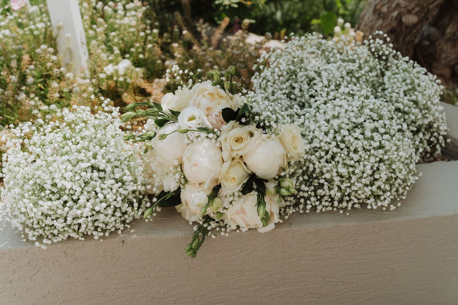 White roses and baby's breath wedding bouquet arrangement on a white surface with lush floral backdrop.