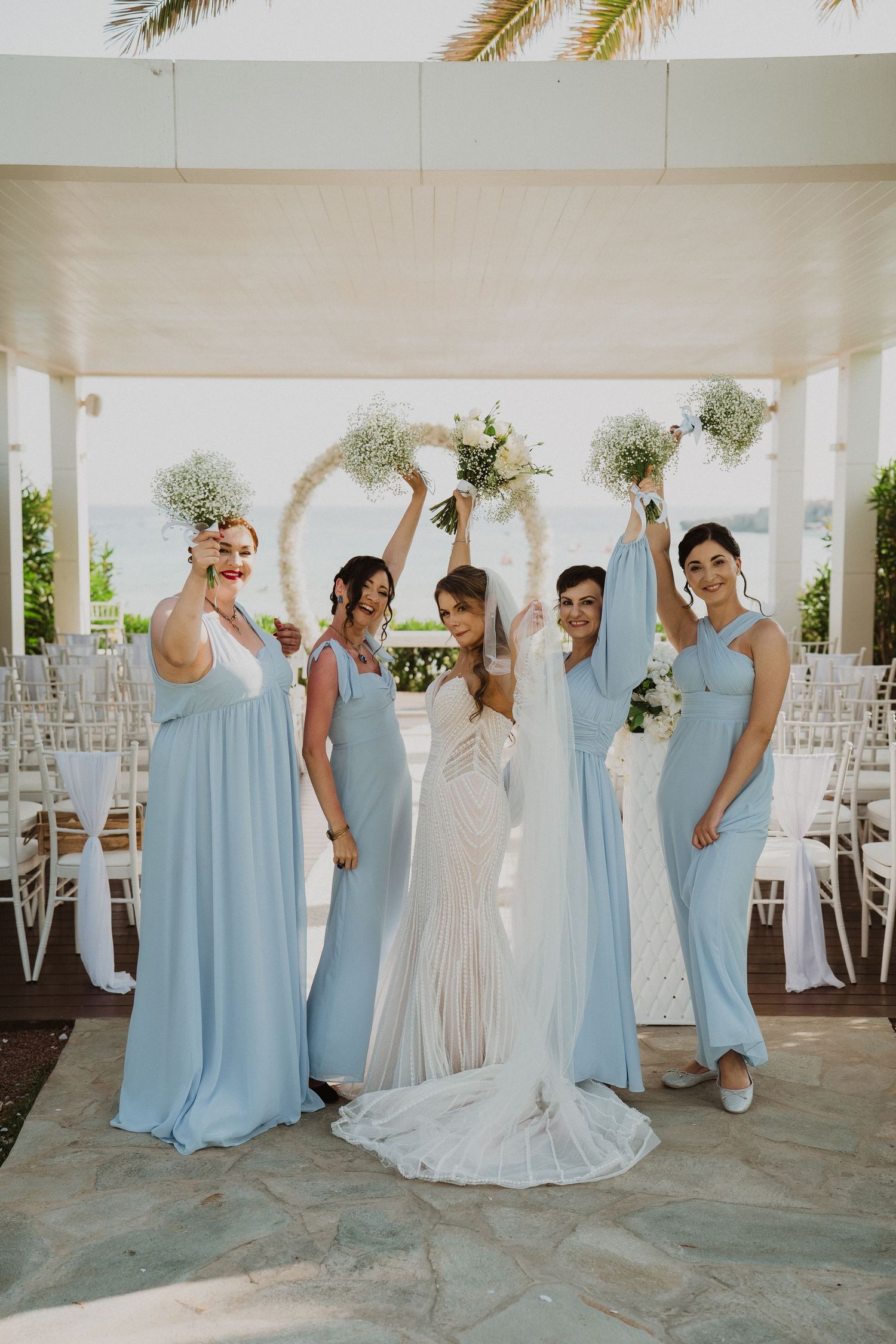 Bride with four bridesmaids in light blue dresses raising bouquets of baby's breath before a circular floral arch.