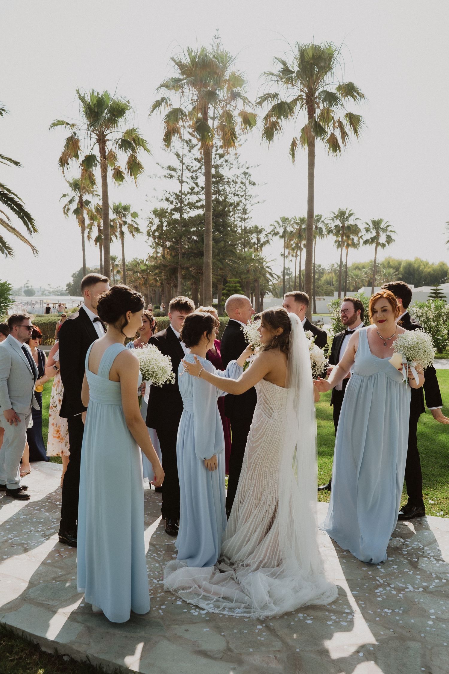 Bride in white gown with bridesmaids in light blue dresses celebrating outdoors with palm trees.