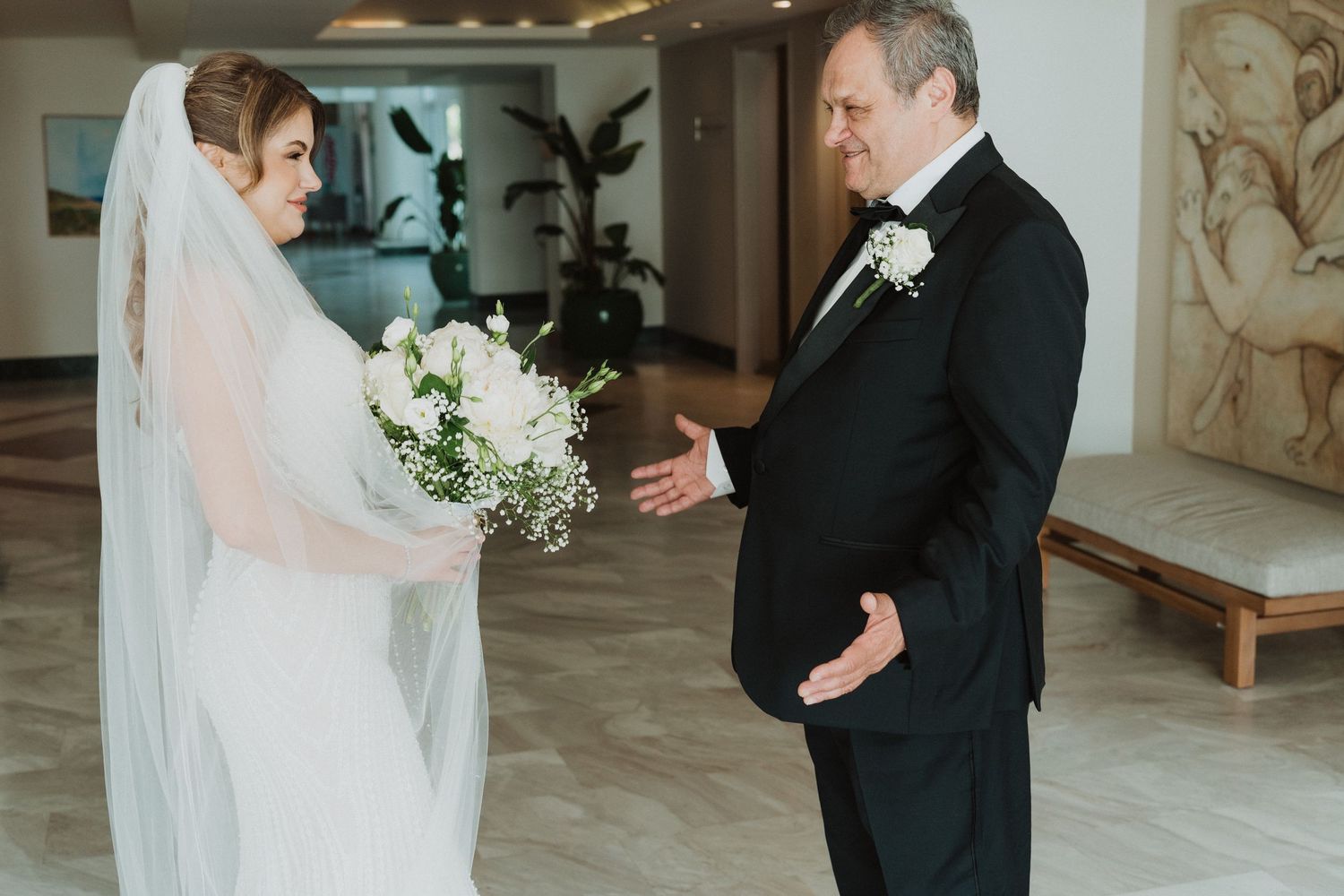Bride in white veil holding bouquet meets older man in tuxedo during wedding first look moment.