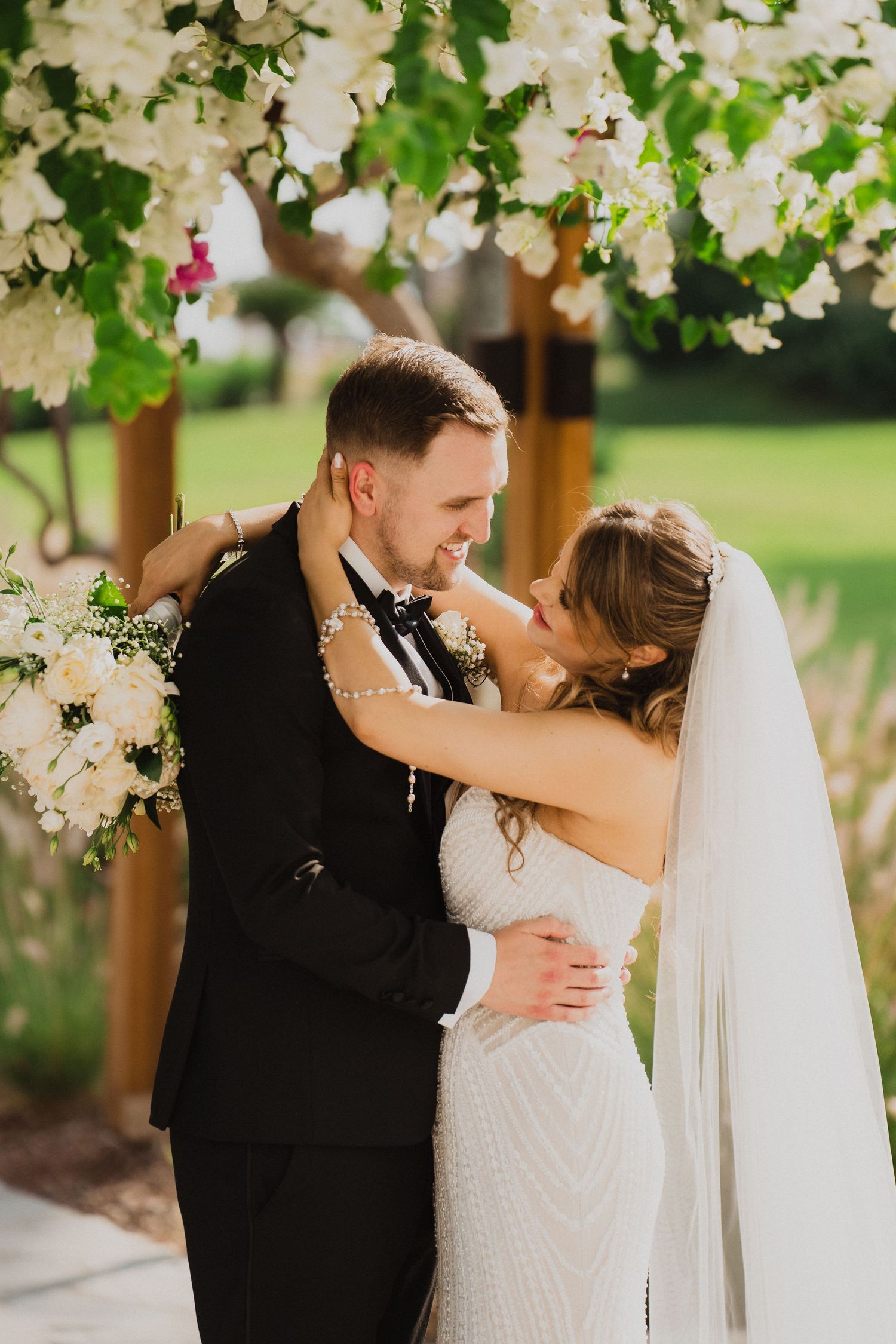 Bride and groom share romantic moment under white floral arch, bride in beaded gown with pearl bracelet and veil.