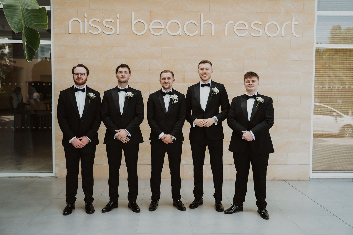 Five groomsmen in black tuxedos with bow ties pose in front of Nissi Beach Resort sign for a wedding photo.