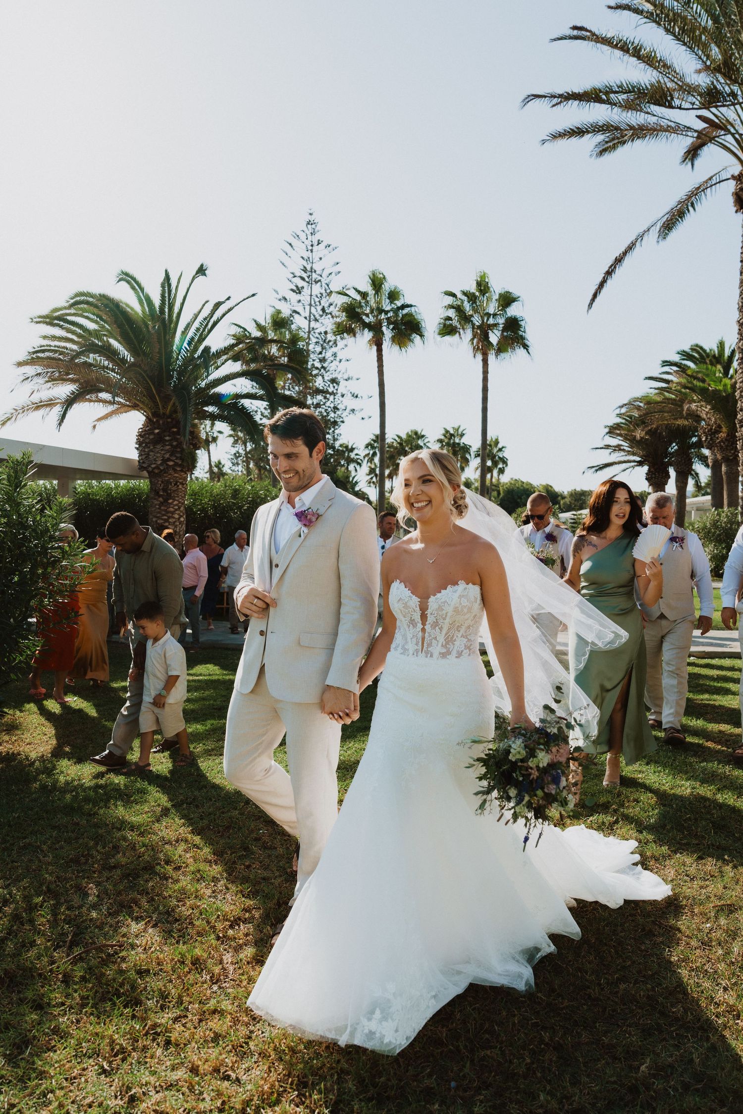 Newlyweds walk through tropical garden setting with palm trees and greenery.