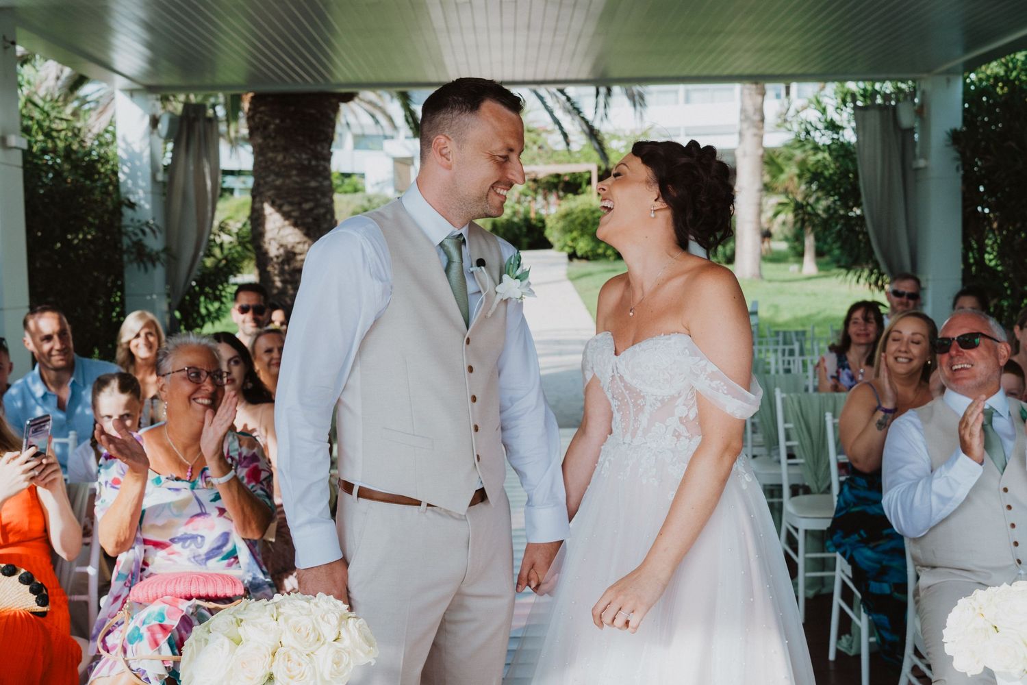 Joyful couple in wedding attire share a moment under a covered porch while guests look on during outdoor ceremony.