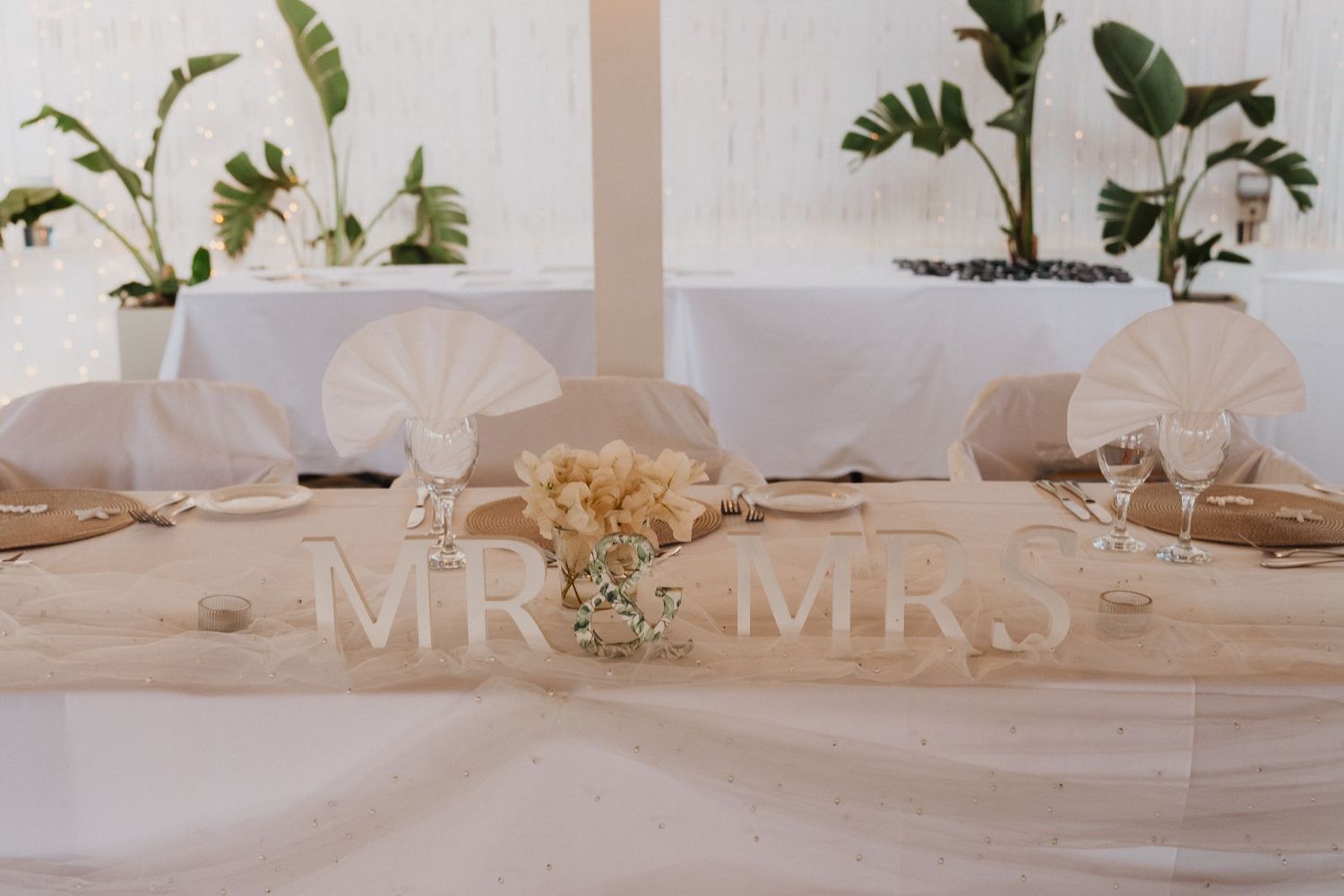 Elegant wedding table setting with 'MR & MRS' sign, neutral linens, and potted plants in white decor.