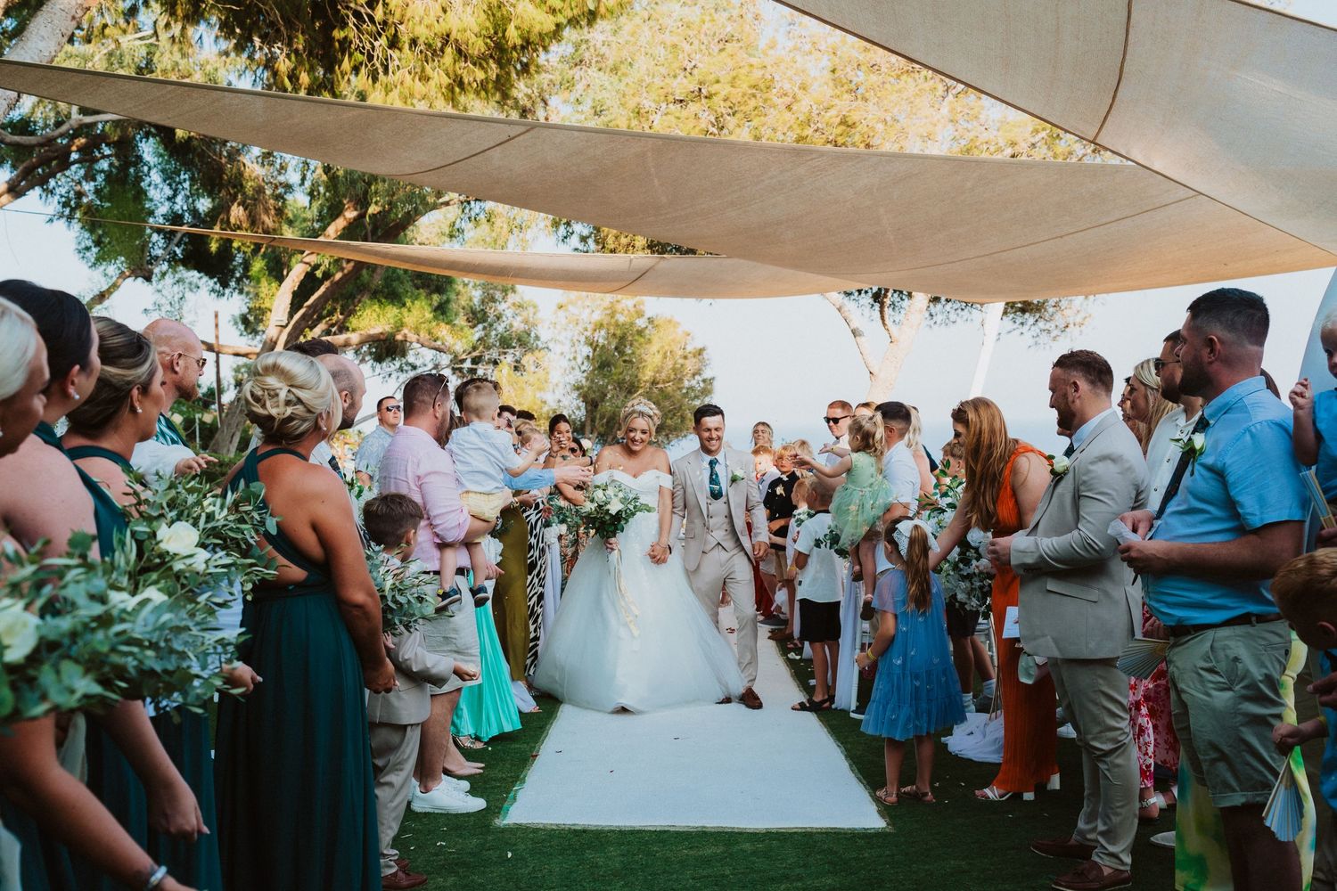 Outdoor wedding ceremony guests standing along a white aisle runner under shade sails.