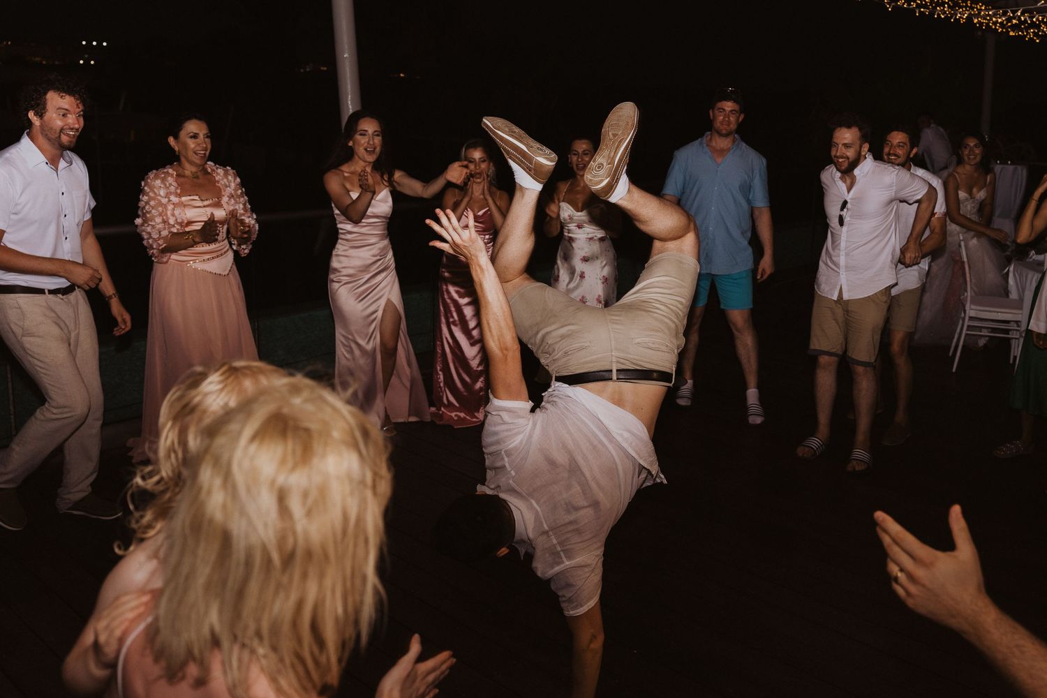 Reception guests cheer as wedding guest performs acrobatic dance move under string lights.
