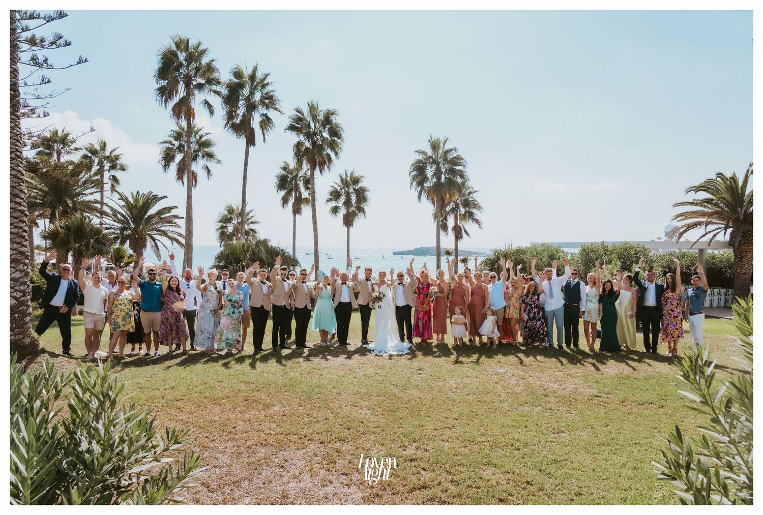 Wedding party guests line up for a group photo on a grassy lawn with tall palm trees.