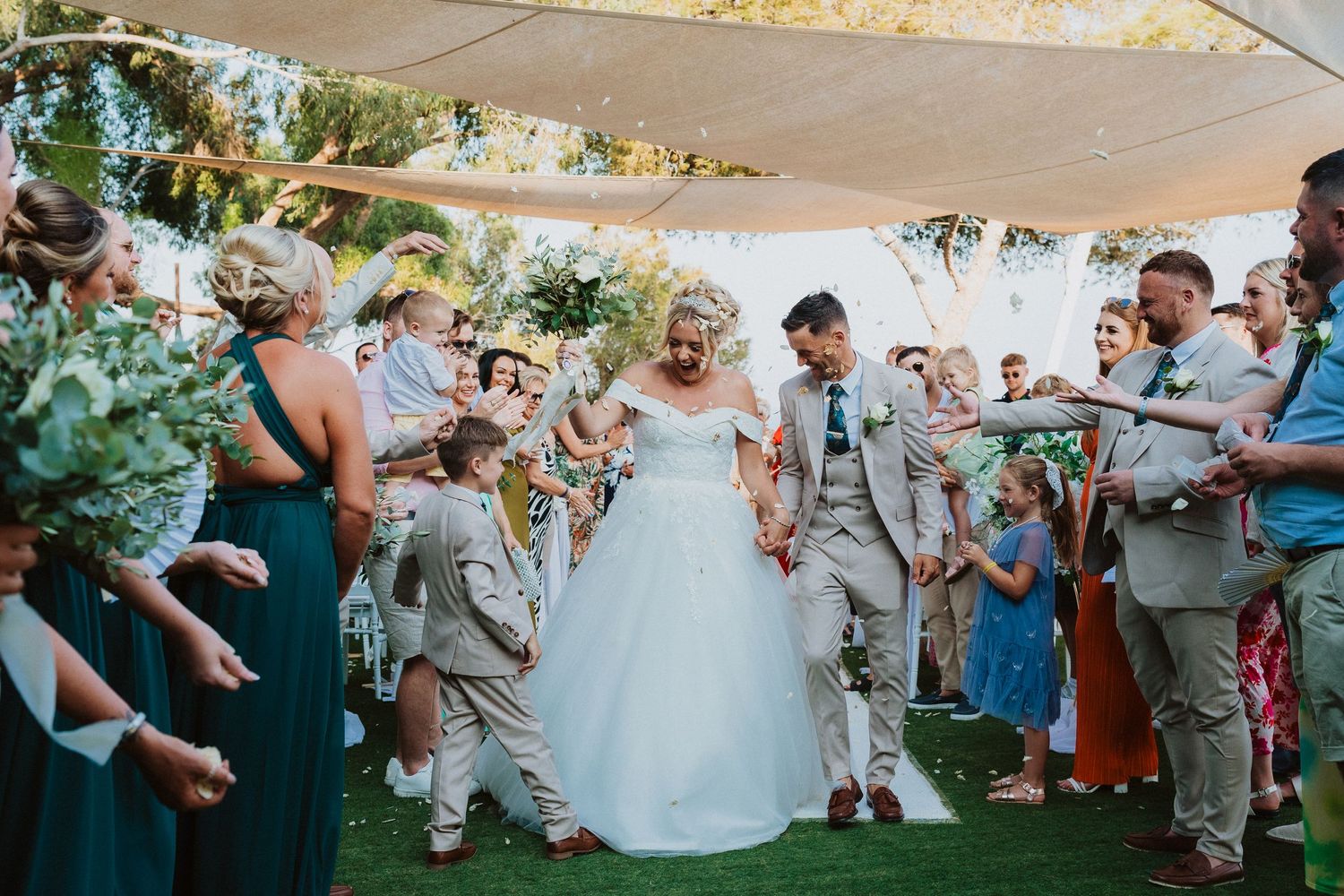 Wedding guests celebrate and take photos as newlyweds walk down an outdoor aisle.