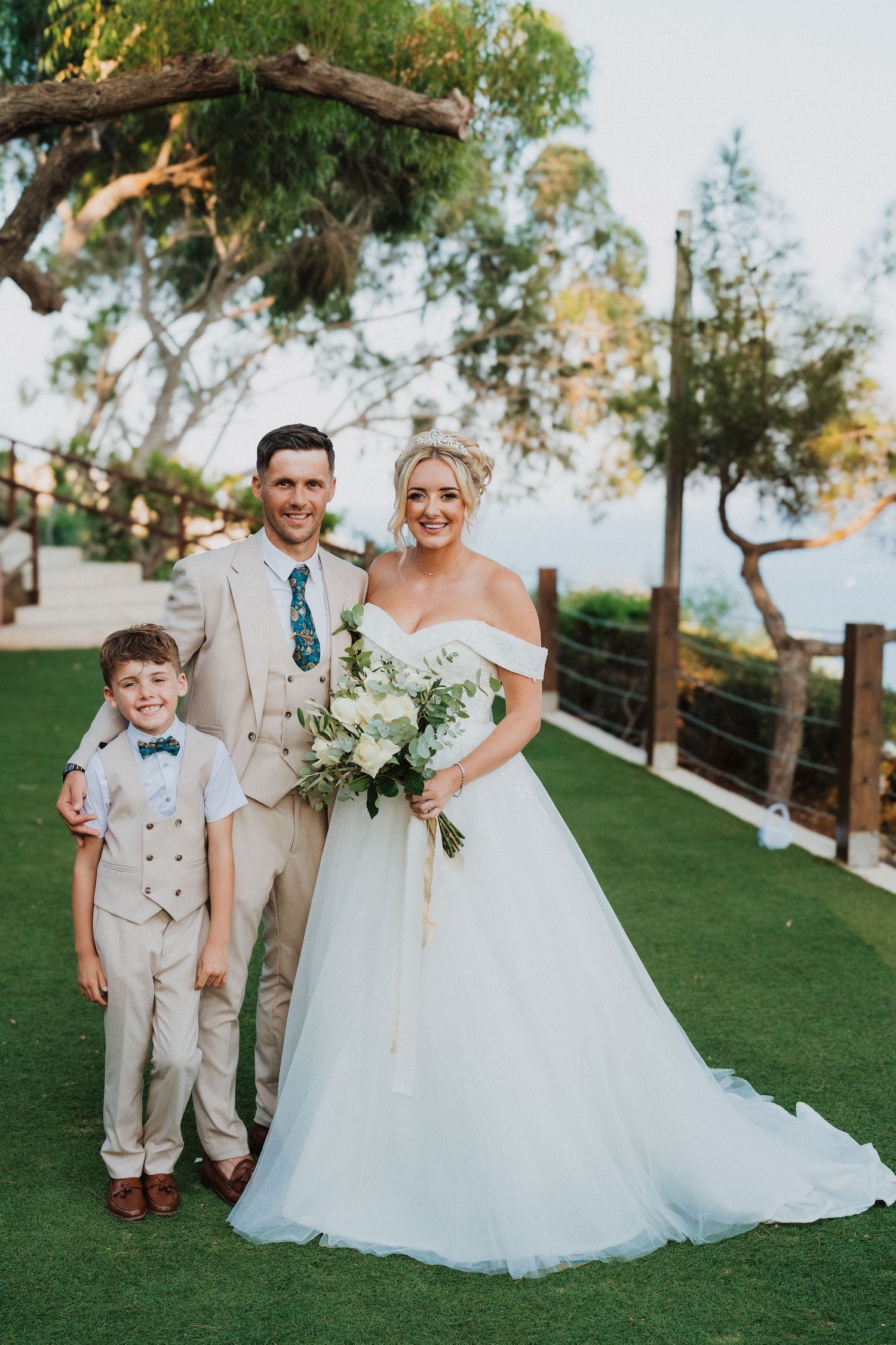 Beautiful outdoor wedding portrait with bride and groom in beige suit with young boy on manicured lawn.
