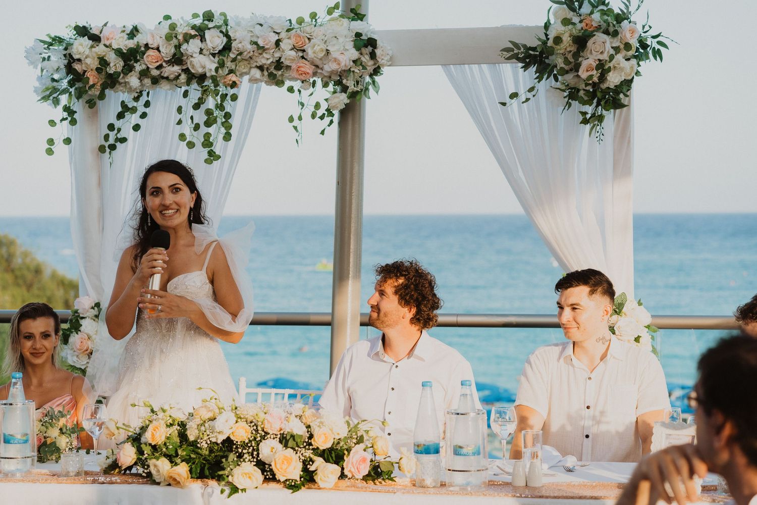 Wedding party seated at decorated dining table with floral arch and ocean backdrop.