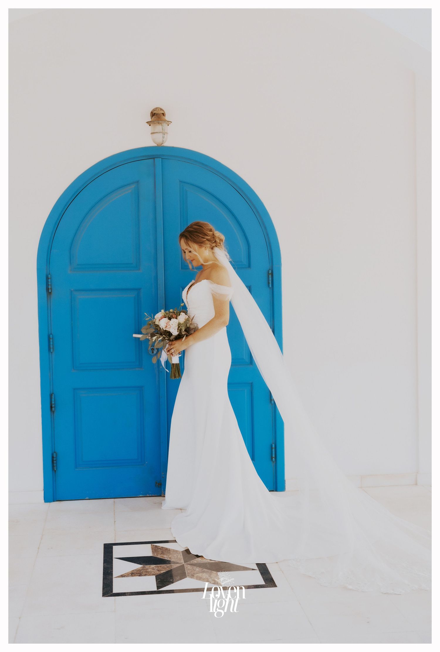 Bride in elegant white gown poses with bouquet against bright blue arched doorway.