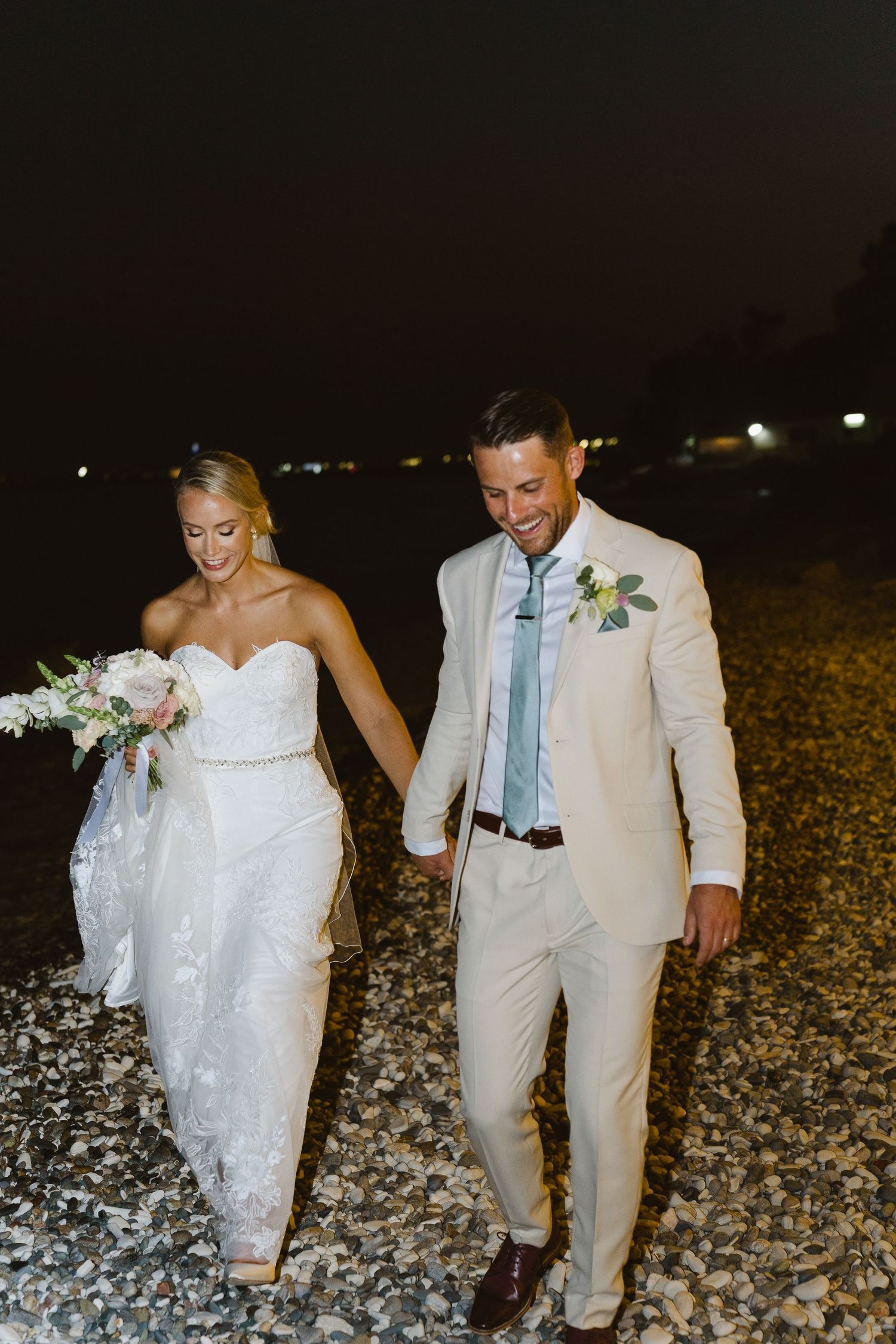Newlyweds walking together on rocky beach during nighttime celebration.