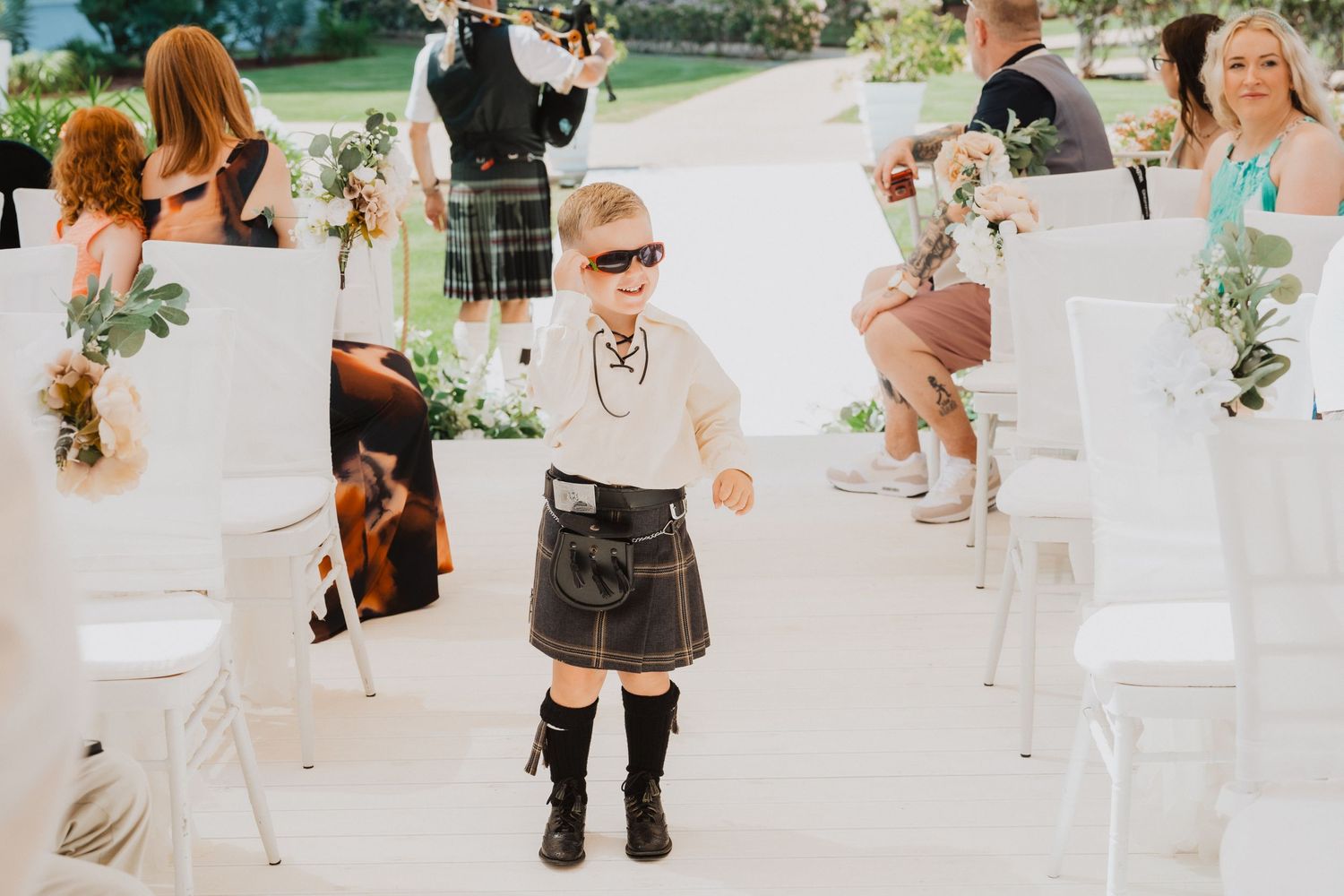 Young ring bearer in a traditional kilt and boots walking between white ceremony chairs.