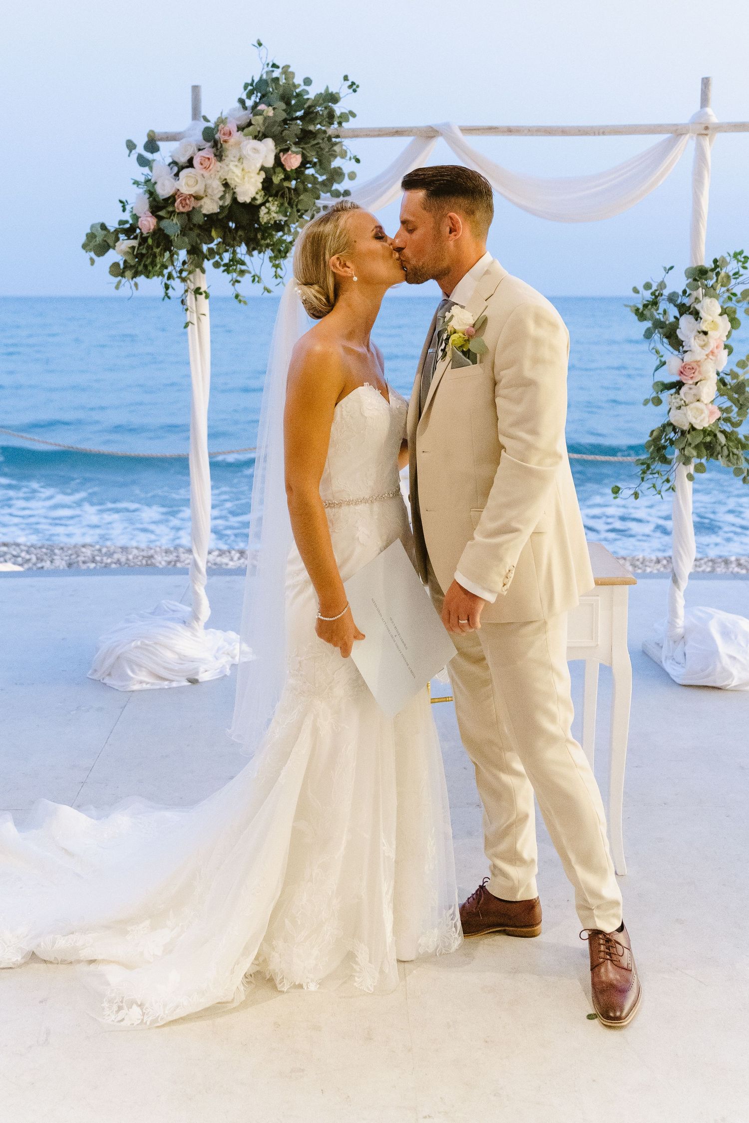 Newlyweds share a kiss under an elegant beach wedding arch decorated with white flowers.
