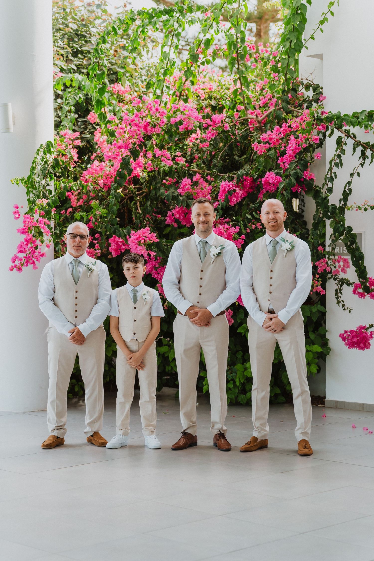 A wedding party wearing cream colored suits stands in front of a vibrant pink bougainvillea bush.