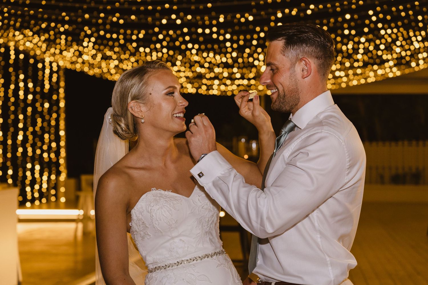 Newlyweds share cake during reception under canopy of golden string lights.