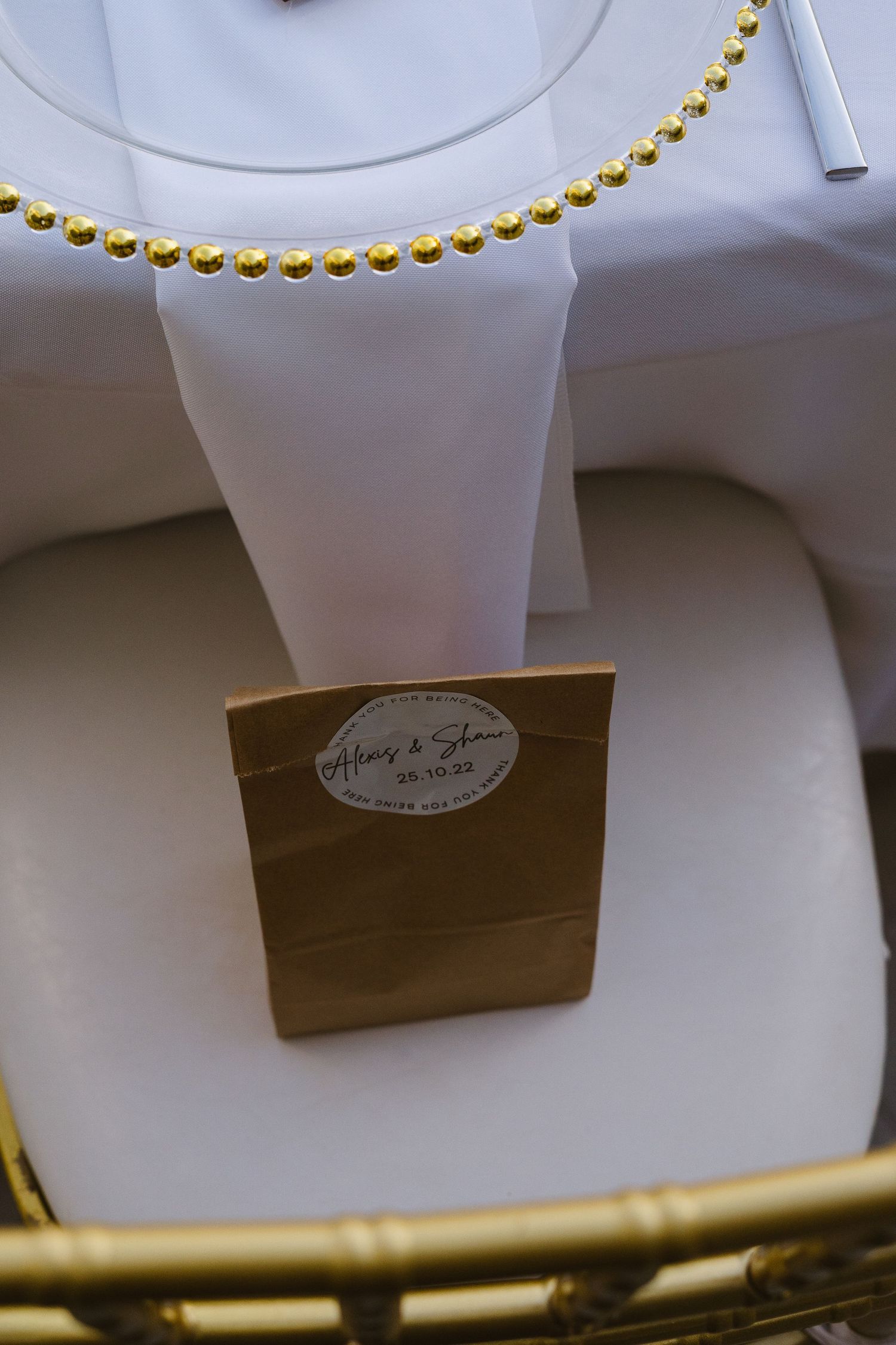Elegant place setting with gold beaded charger plate and brown paper favor bag.