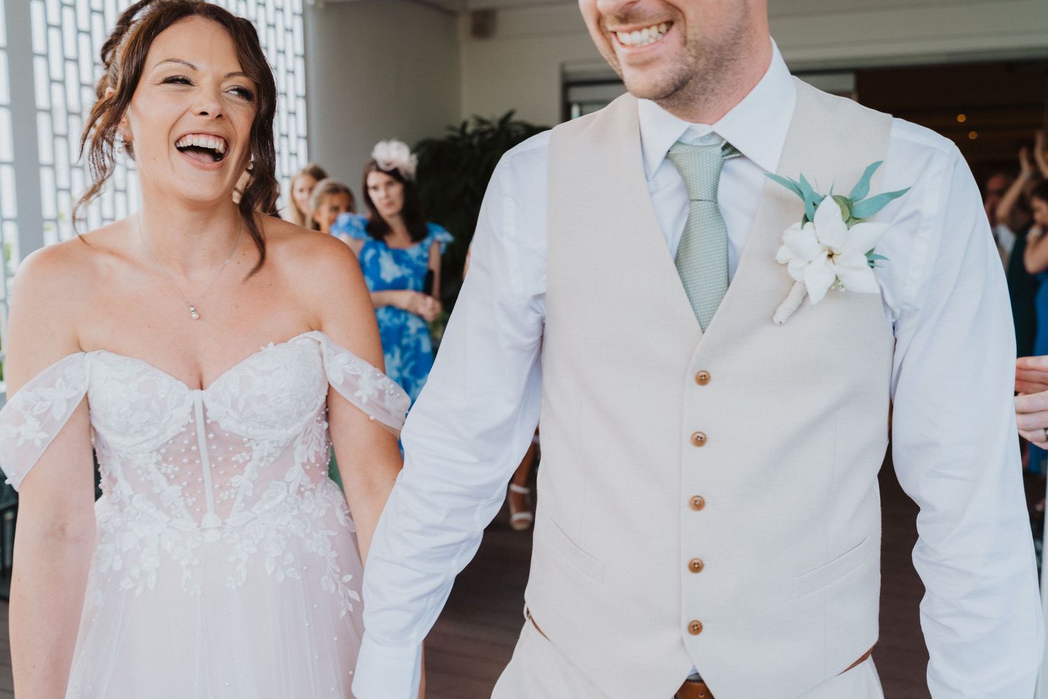 A joyful couple at their wedding celebration, with the bride in a strapless white dress and groom in light vest and tie.