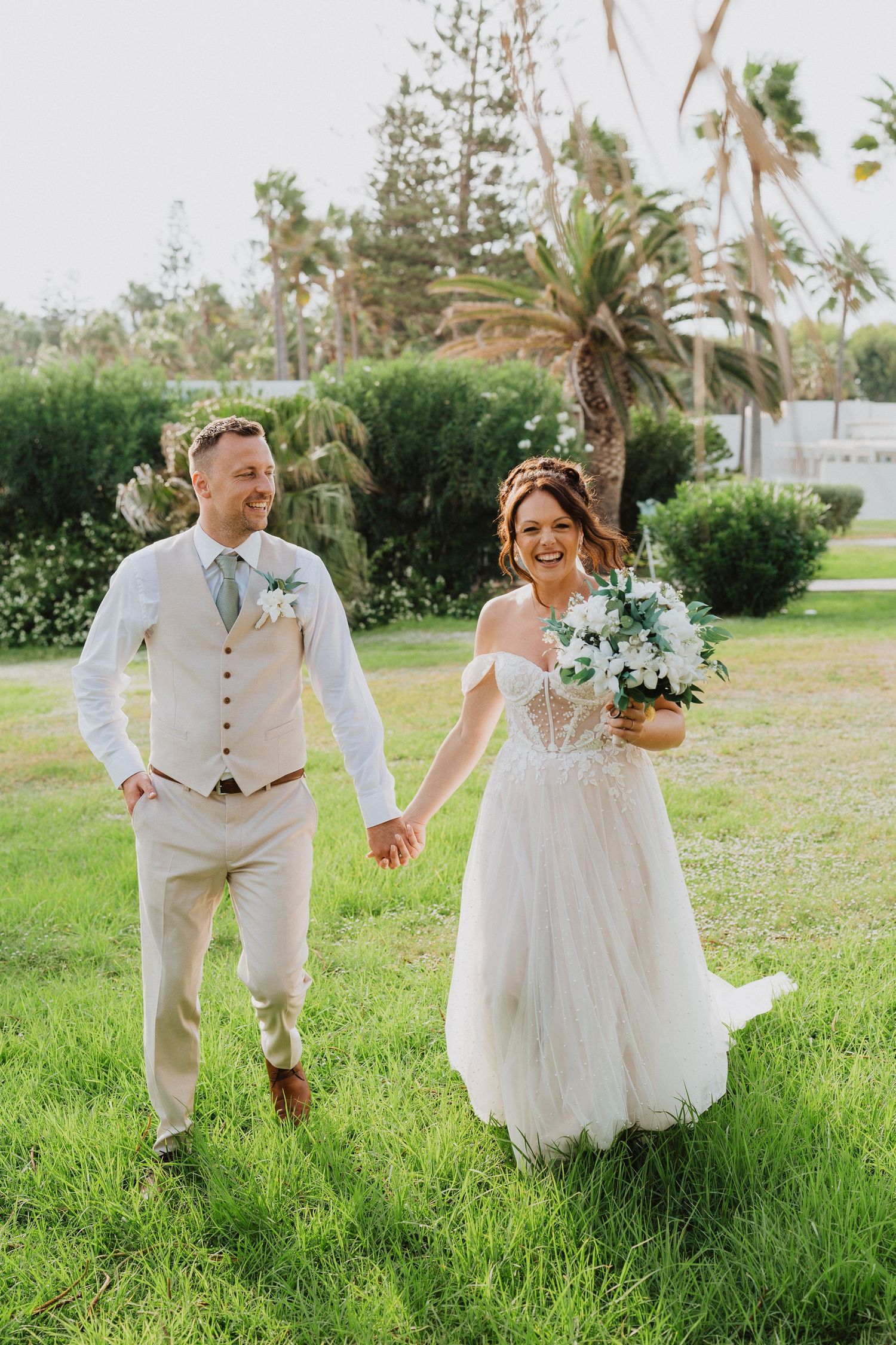 A couple walks hand in hand on green grass at a tropical wedding venue with palm trees in the background.