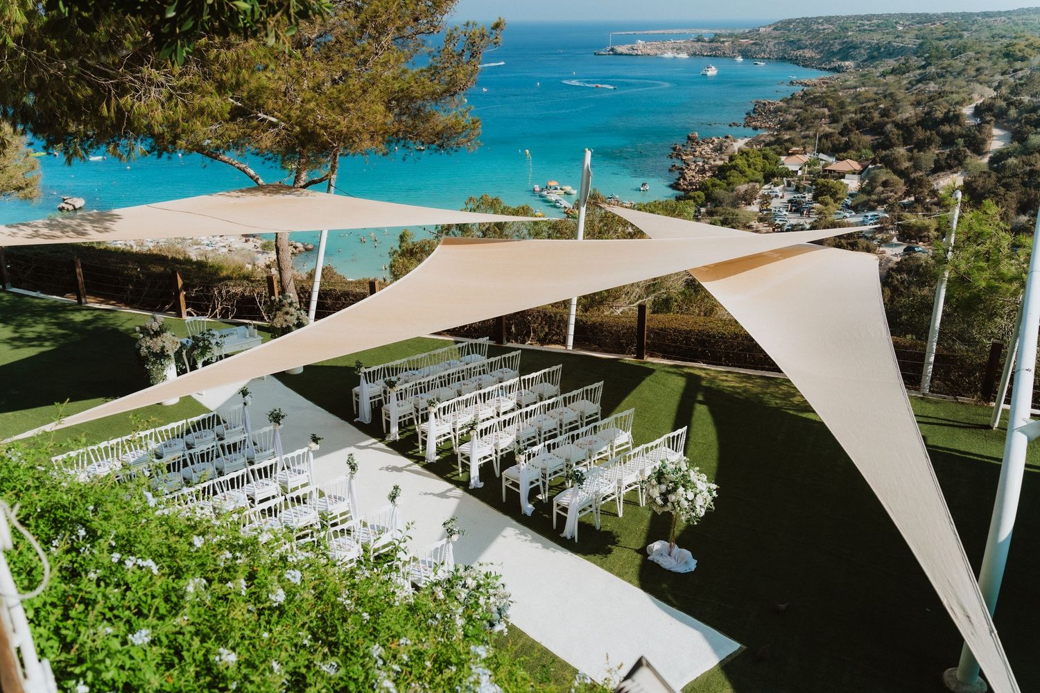 Stunning outdoor wedding ceremony setup with white chairs and sail shades overlooking a bright blue ocean bay.