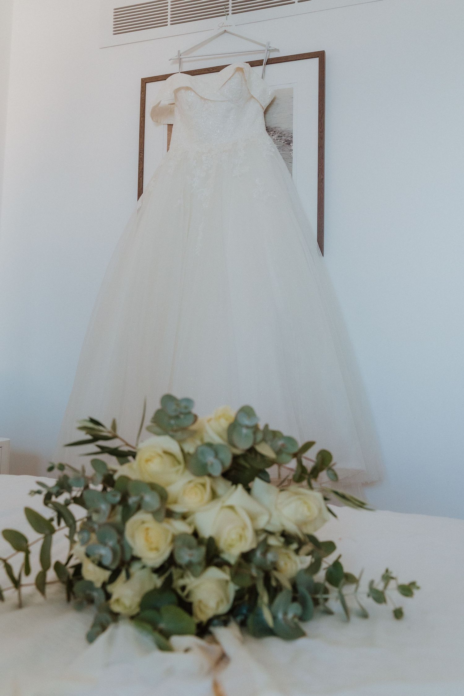White wedding dress hangs above elegant floral arrangement with cream roses and eucalyptus leaves on white surface.
