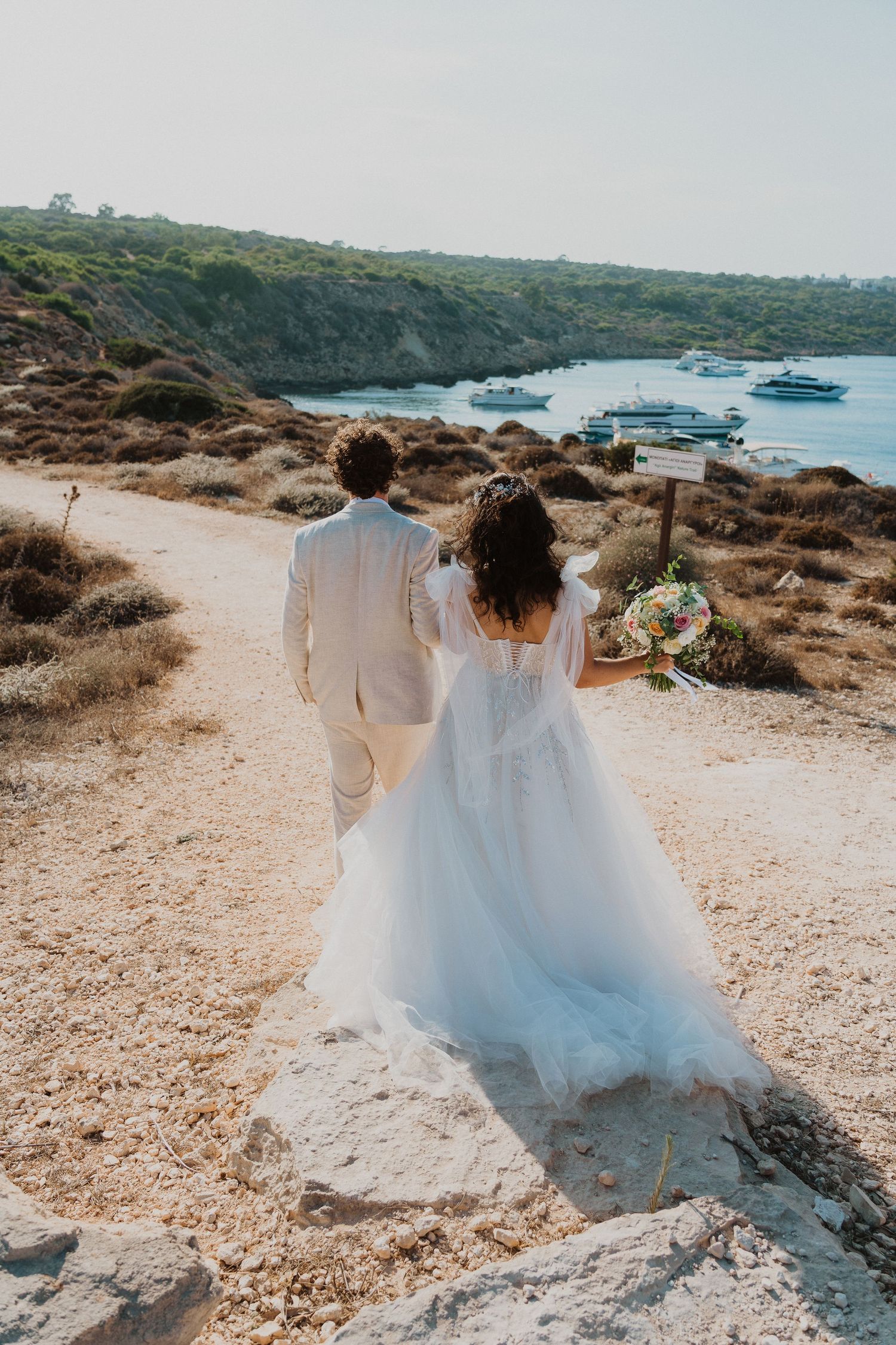 A couple in wedding attire walk along a coastal path overlooking boats anchored in a Mediterranean cove.