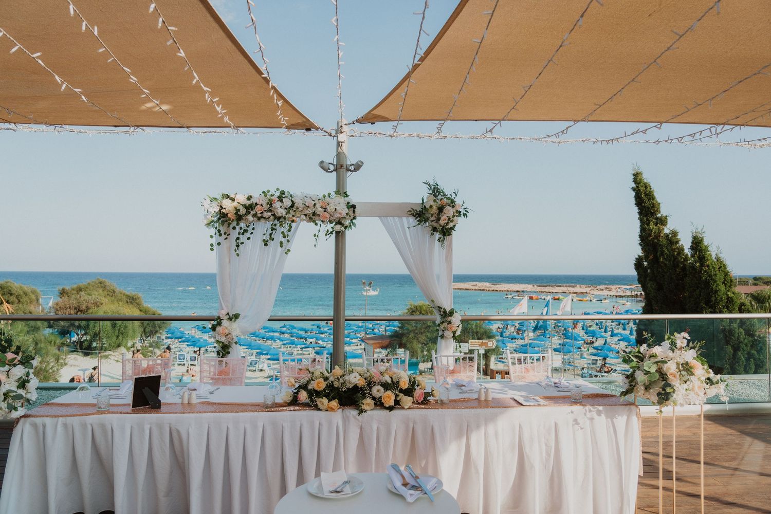 Romantic oceanfront wedding setup with white draping, floral arch and covered dining tables.