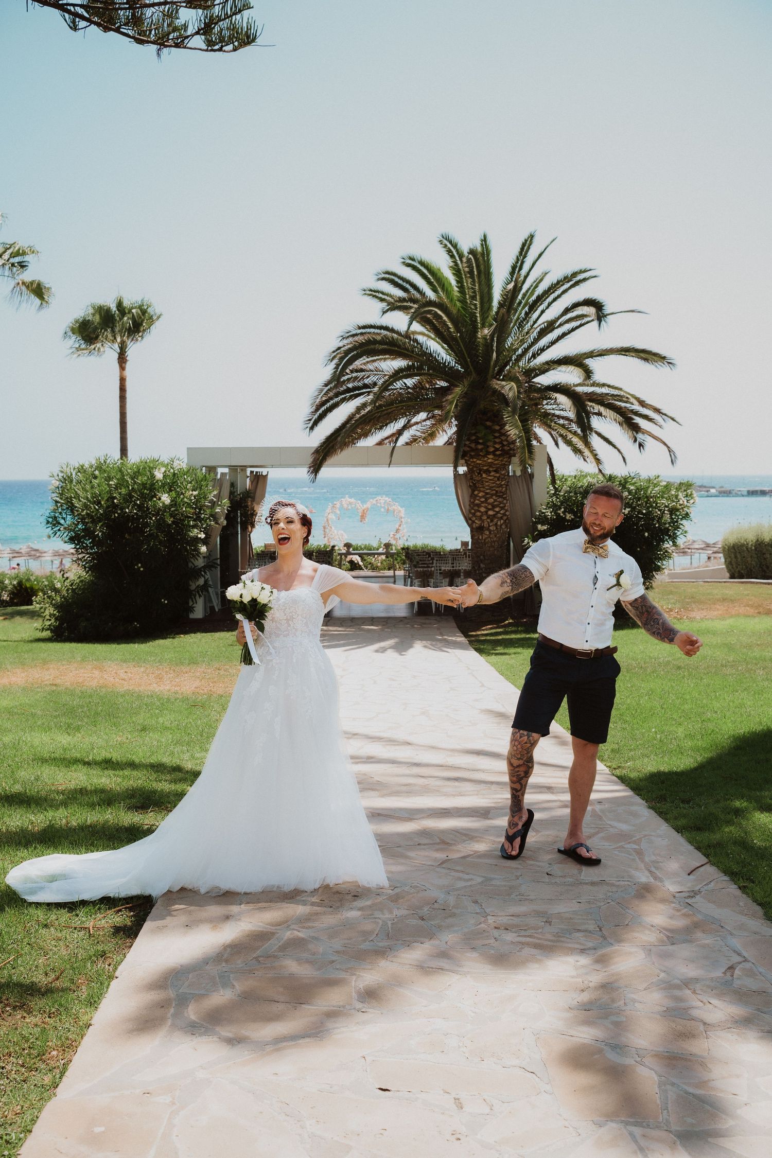 Newlyweds share a joyful moment on a seaside path with swaying palm trees overhead.
