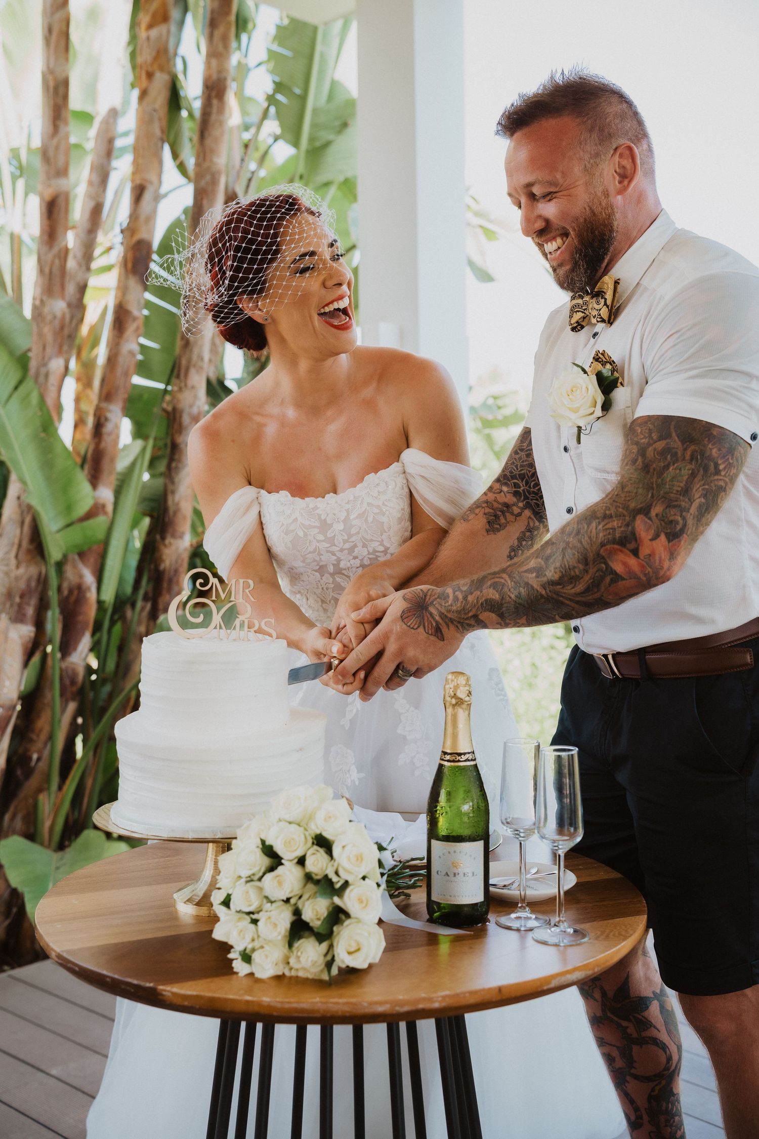 Joyful couple laughing while cutting their wedding cake on wooden table.