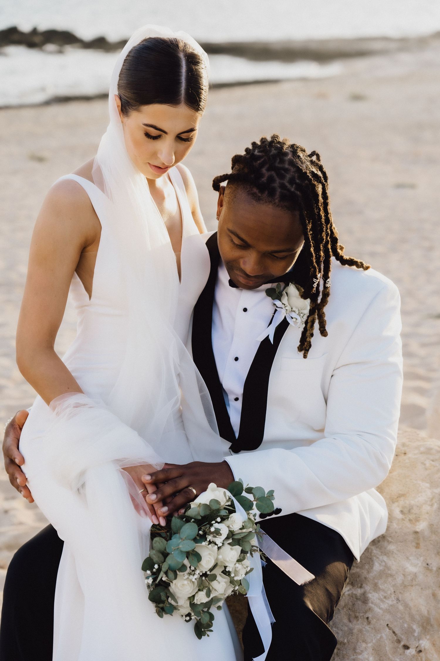 Elegant couple in white wedding attire share tender moment with bouquet on beach.
