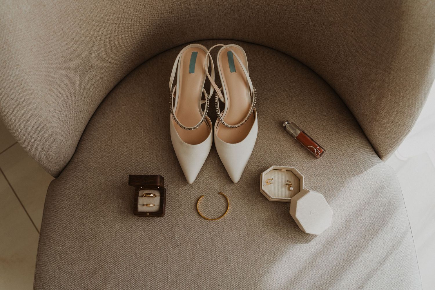 Wedding accessories and white heels arranged on beige upholstery with rings and perfume.