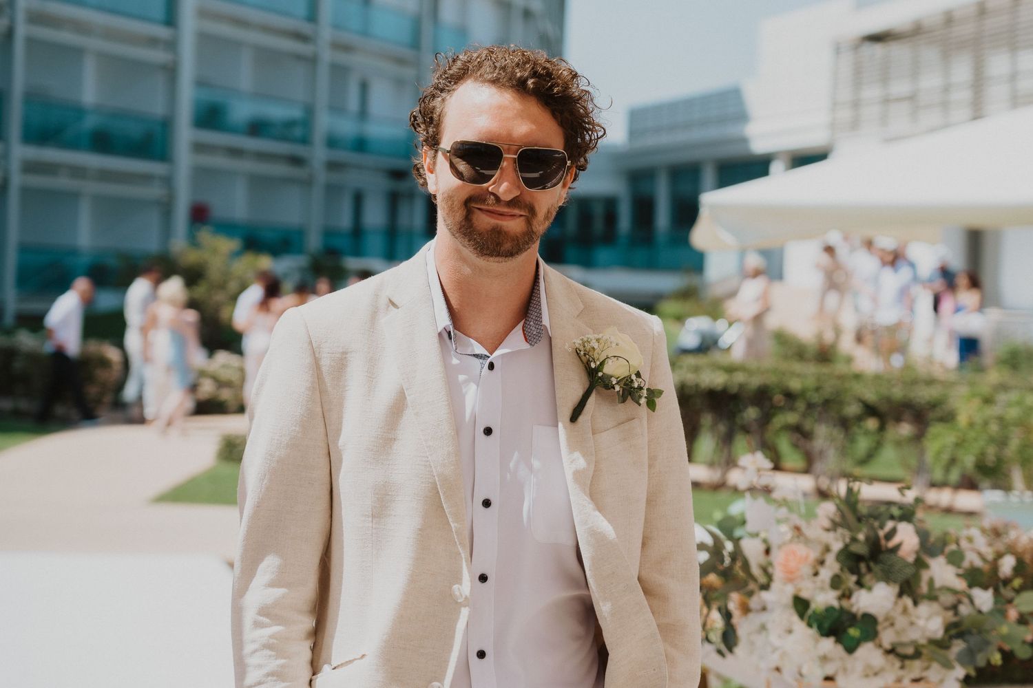 A wedding guest in a cream blazer and sunglasses stands outside a modern venue on a sunny day.