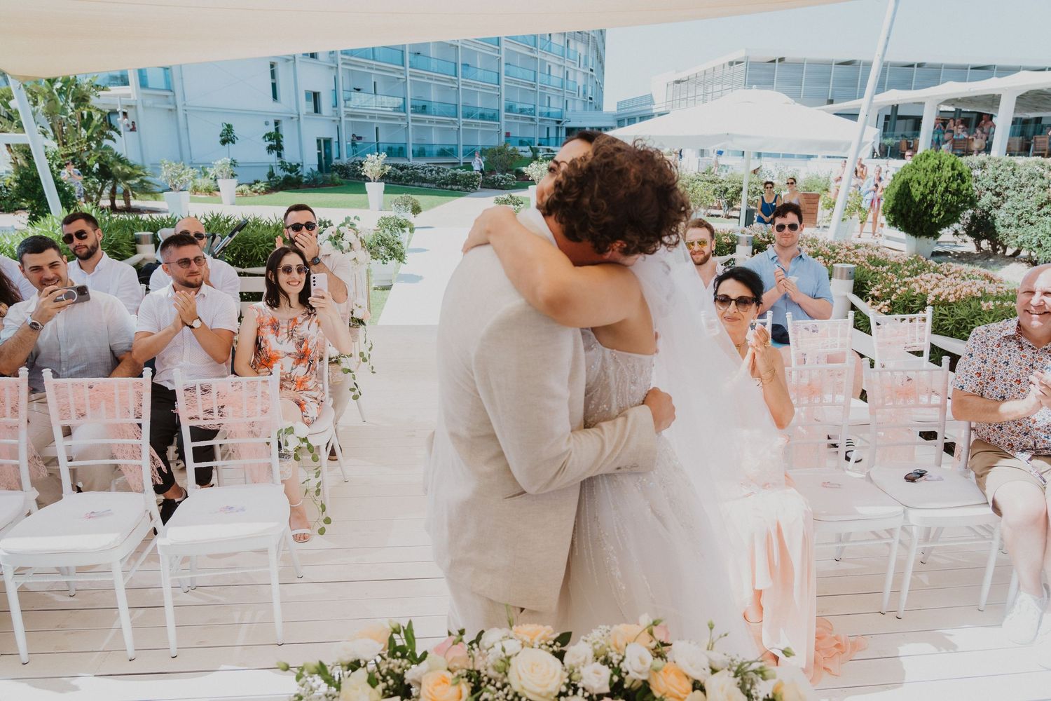 Romantic first kiss at wedding ceremony with guests celebrating in background.
