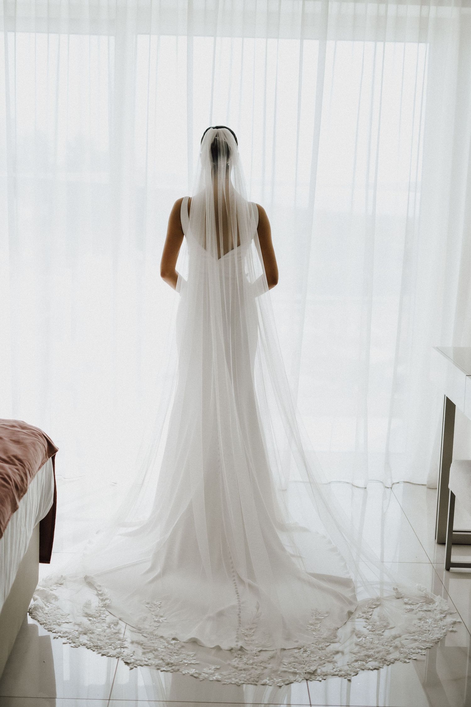 Back view of a wedding dress with long lace-trimmed train and veil against white curtains.