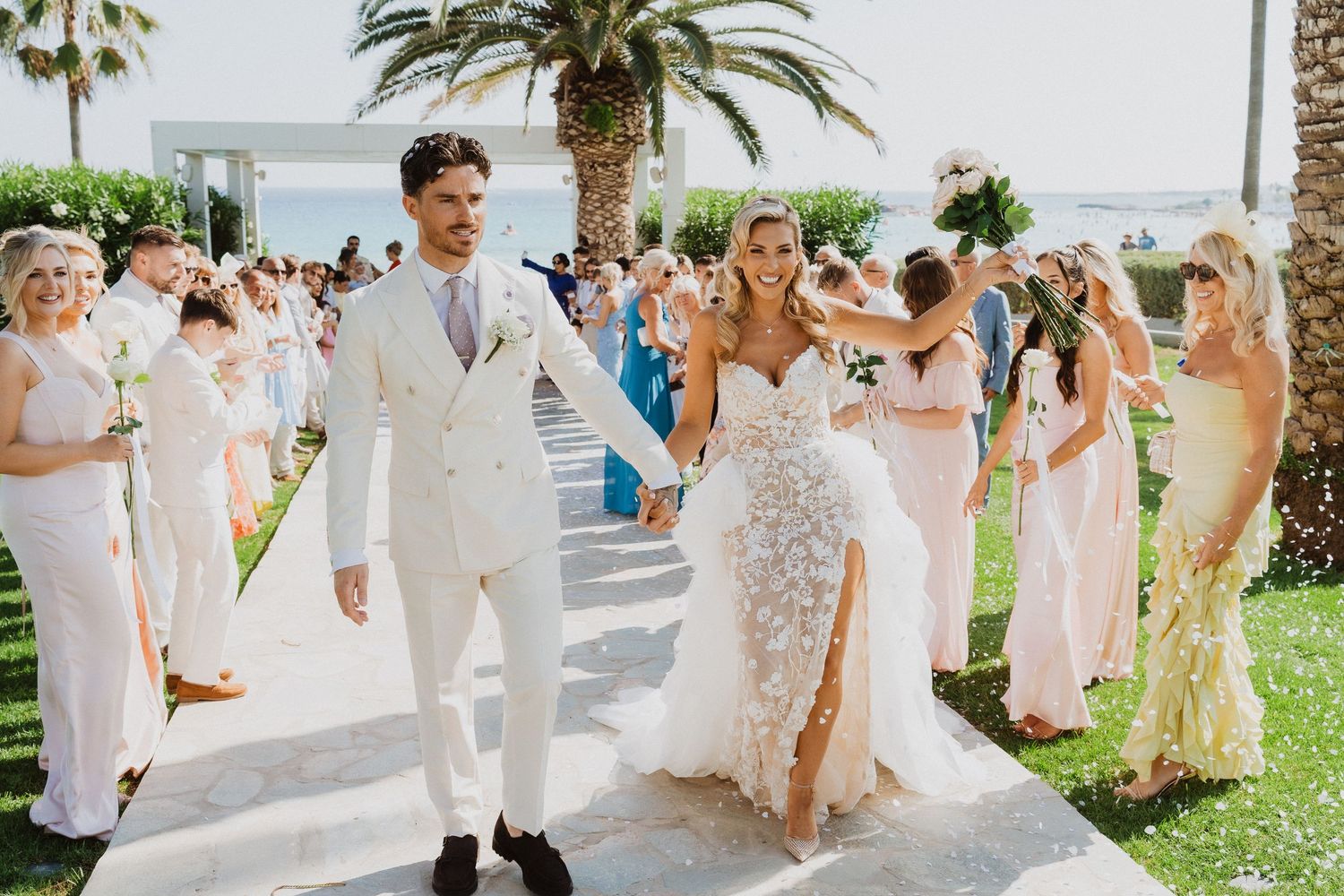Newlyweds walk down aisle lined with guests at outdoor beach wedding ceremony with palm trees in background.