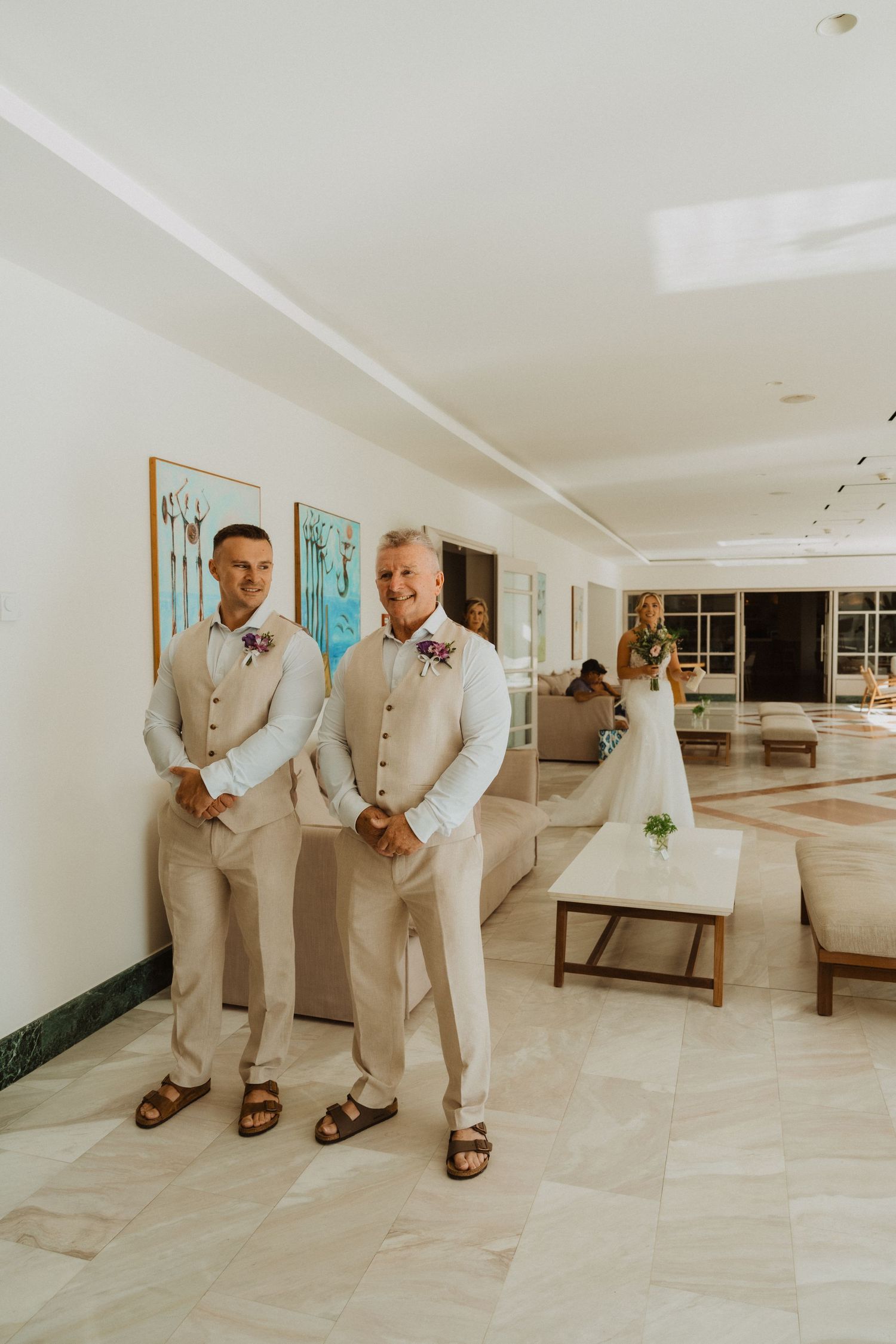 Wedding party members in light colored suits stand casually in a bright indoor space.