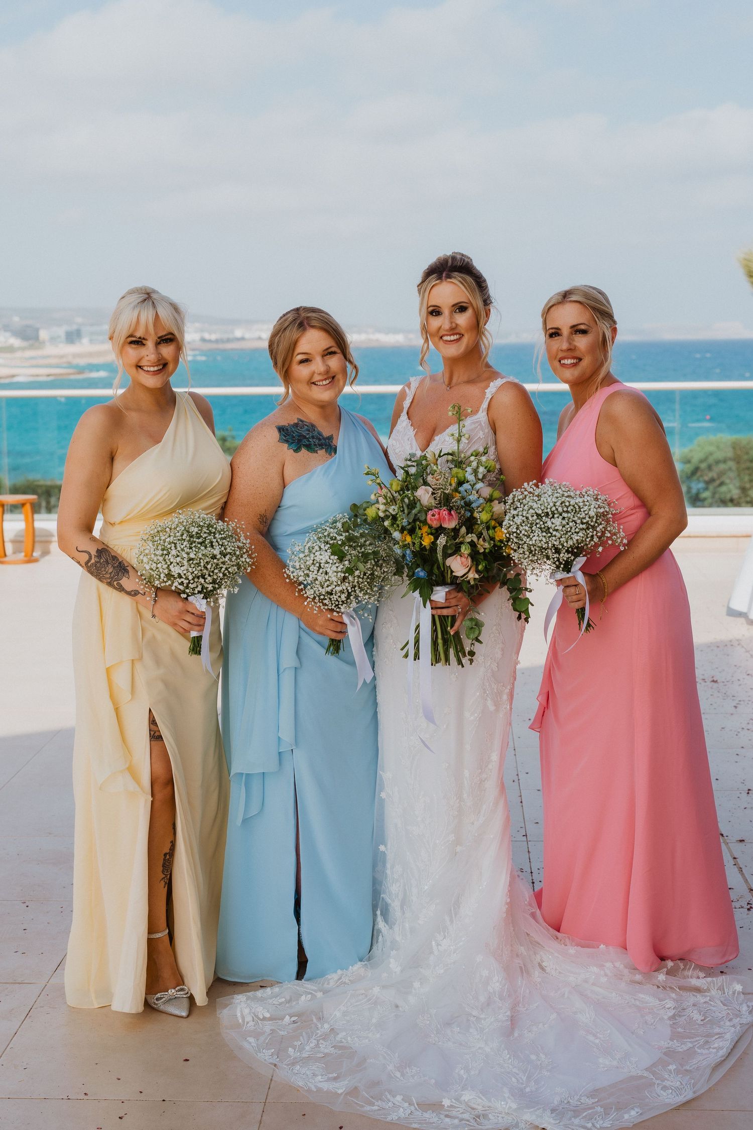 Bridesmaids in pastel yellow, blue and pink dresses pose with bouquets by the ocean on a sunny wedding day.