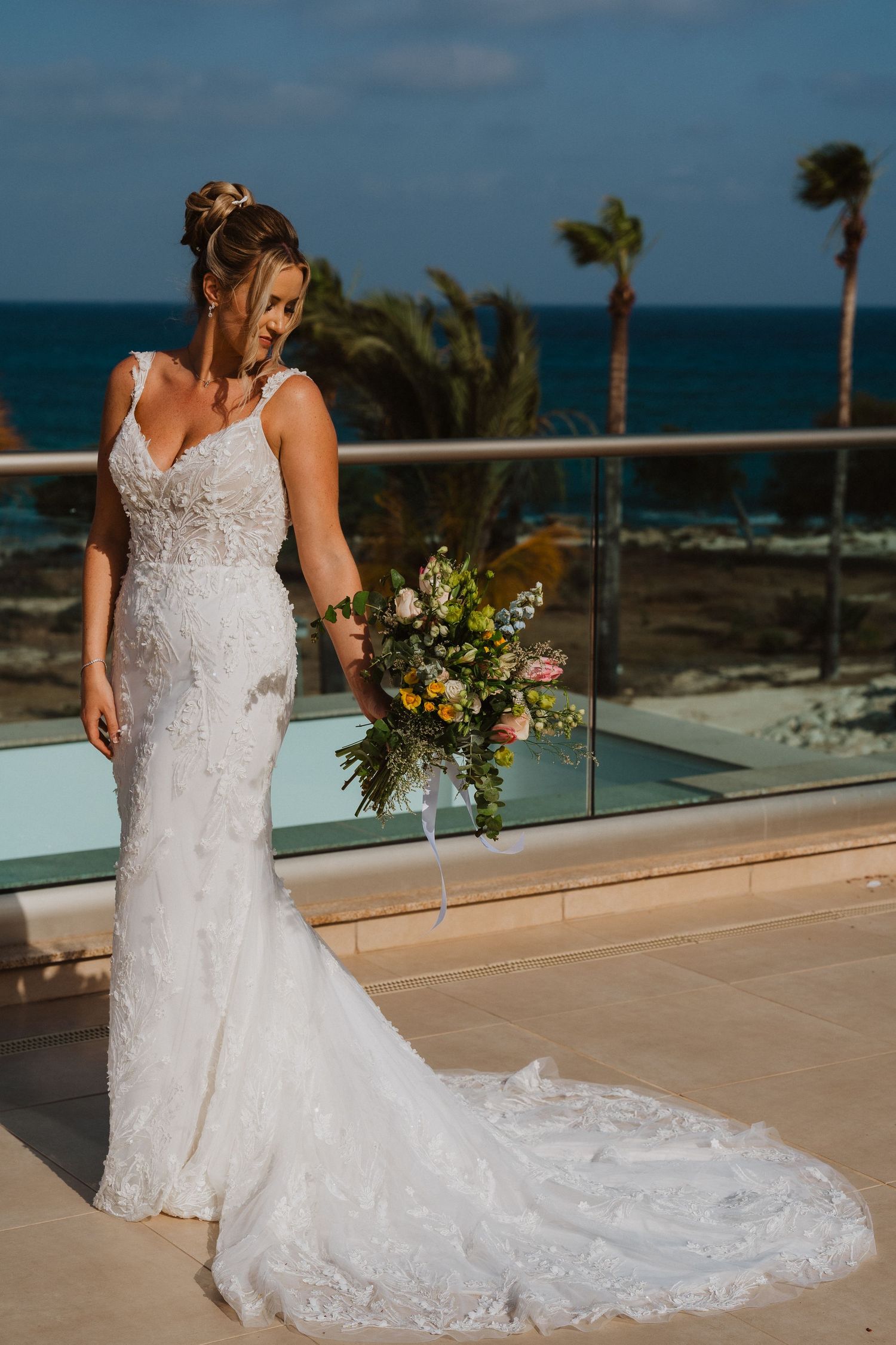 Elegant white lace wedding dress with long train shown from multiple angles on oceanfront balcony with palm trees.