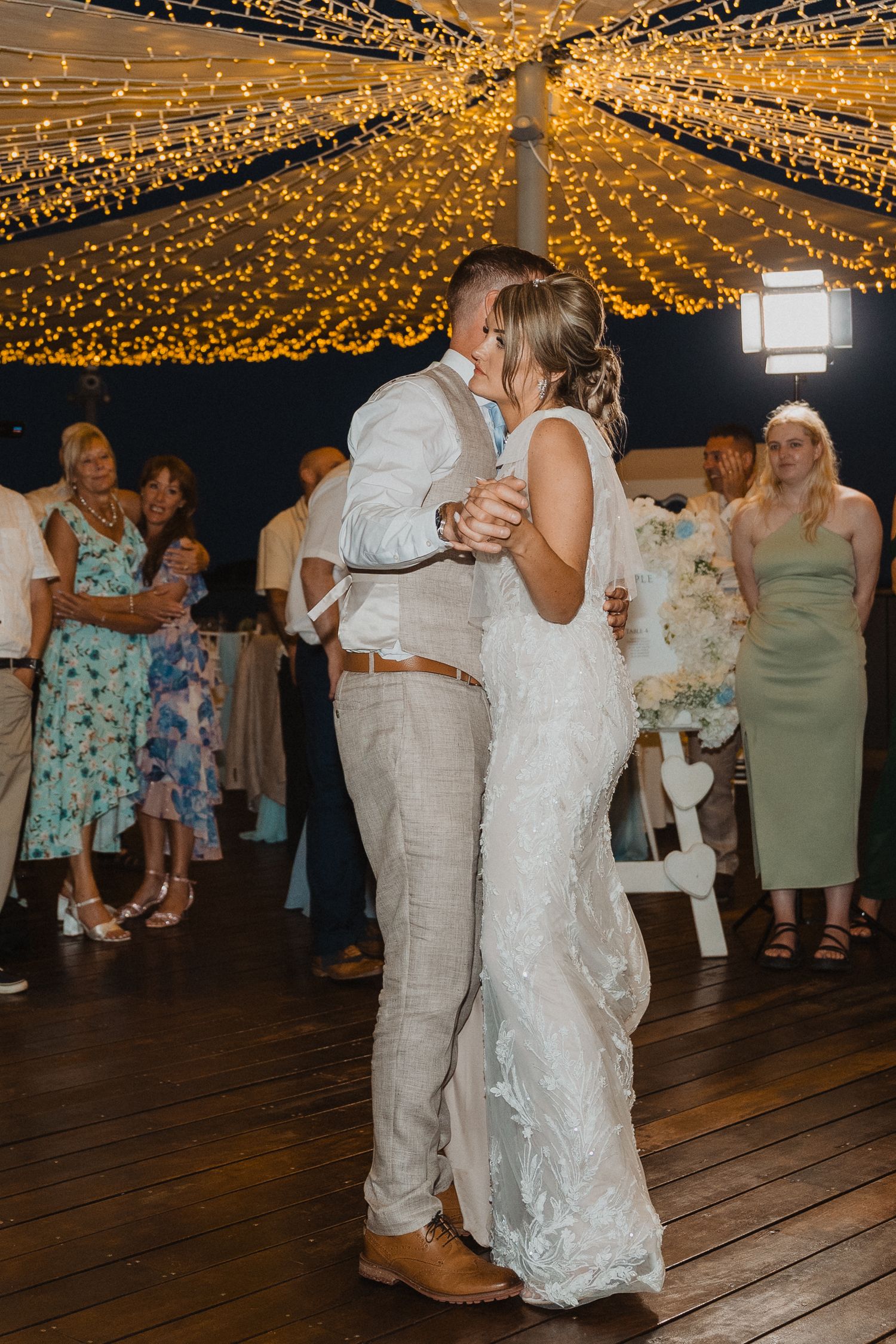 A romantic first dance under twinkling golden lights at a wedding reception with guests watching in the background.