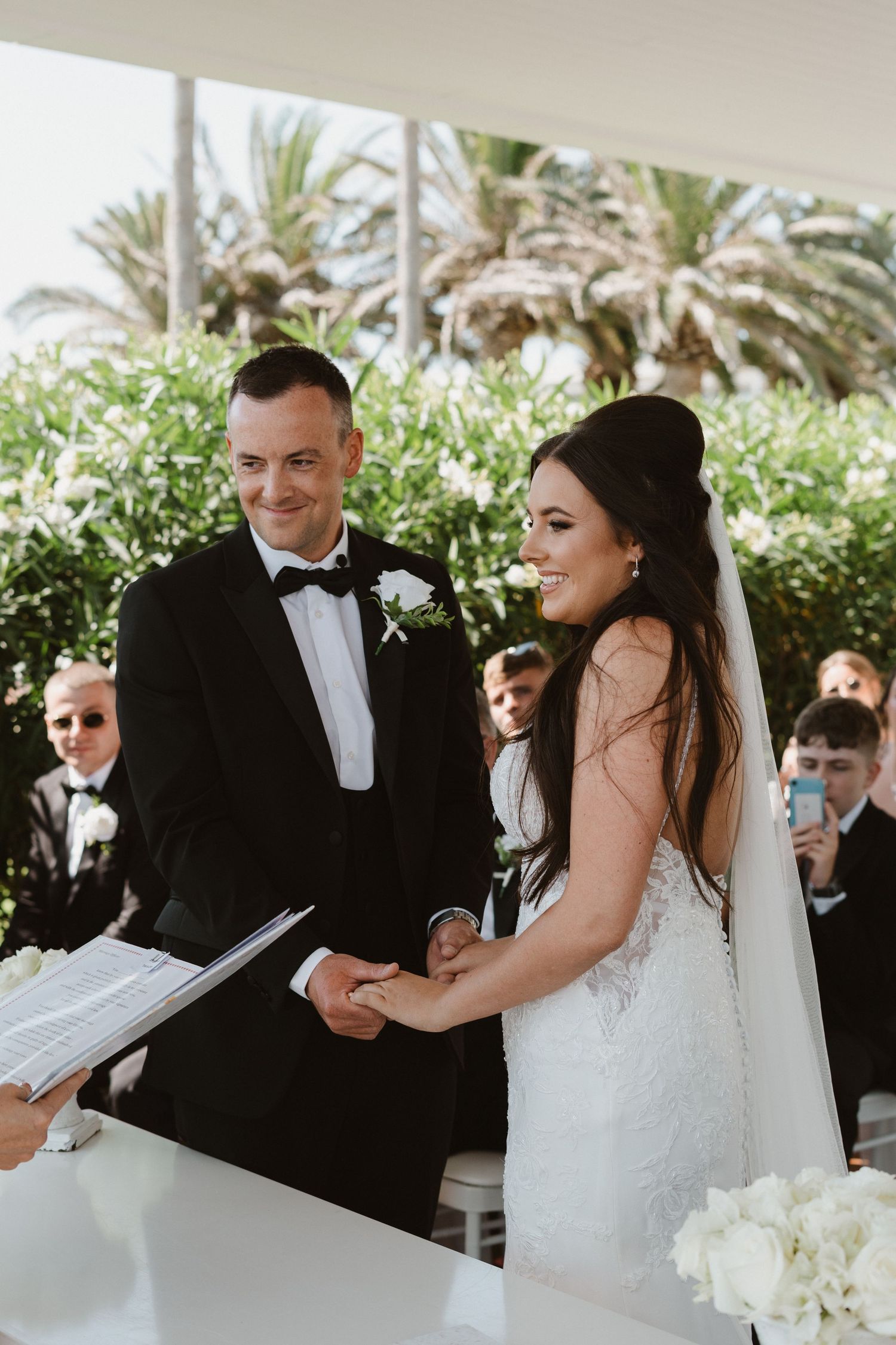 Wedding couple exchanges vows under a white covered patio surrounded by tropical palm trees.