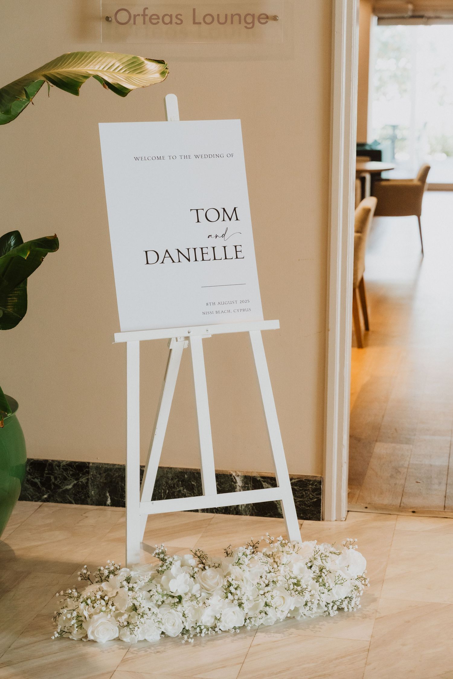 White wedding seating chart signs with floral arrangements displayed on easels in a light-filled venue space.