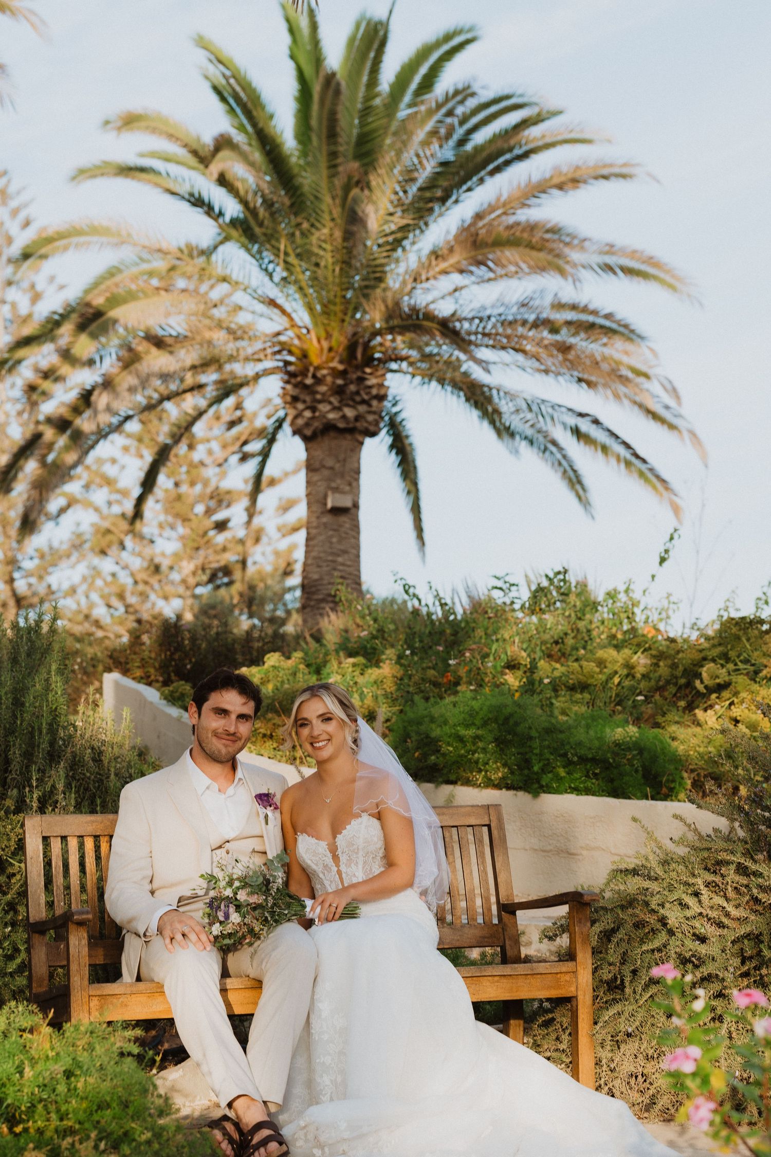 Newlyweds share laughs on wooden bench under palm trees in garden setting.