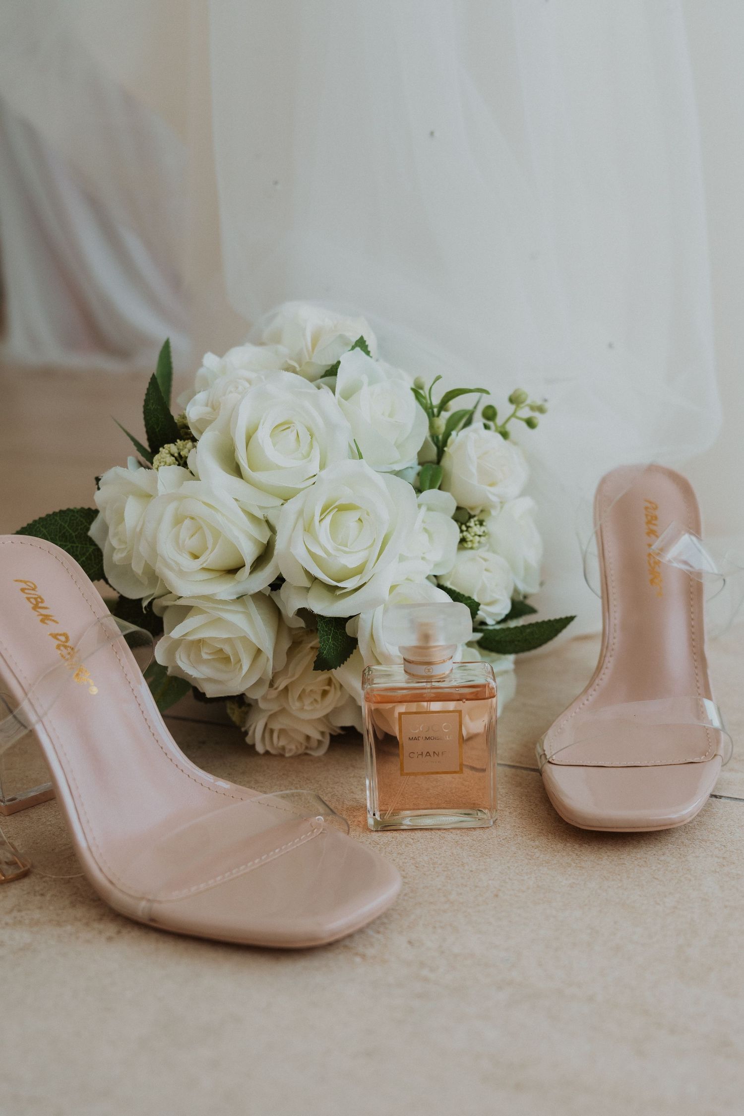 Bridal flat lay with nude clear heeled sandals, white rose bouquet, and amber perfume bottle on beige surface.