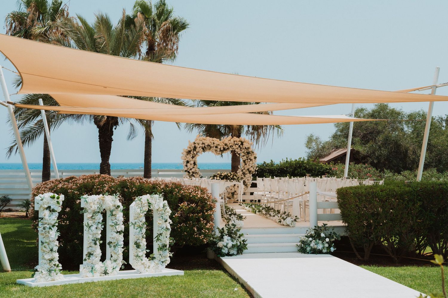 Elegant outdoor wedding ceremony setup with floral arch, number decorations, white aisle, and beige shade sails near the ocean.