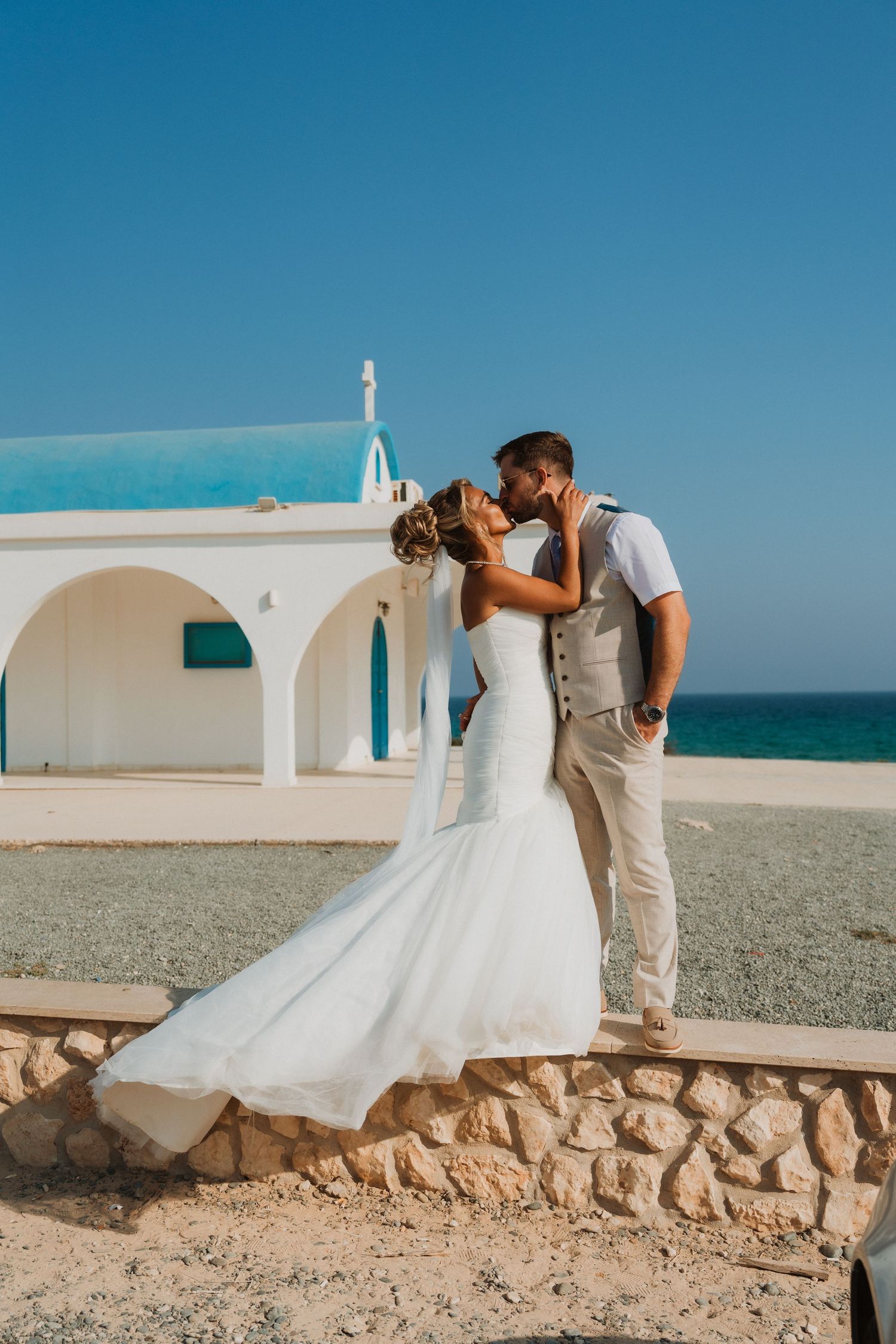 Bride and groom sharing a romantic kiss by a white chapel with turquoise dome near the sea on their wedding day.